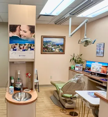 Interior of a dental office with a green dental chair, overhead light, sink with hygiene supplies, and computer on a counter.