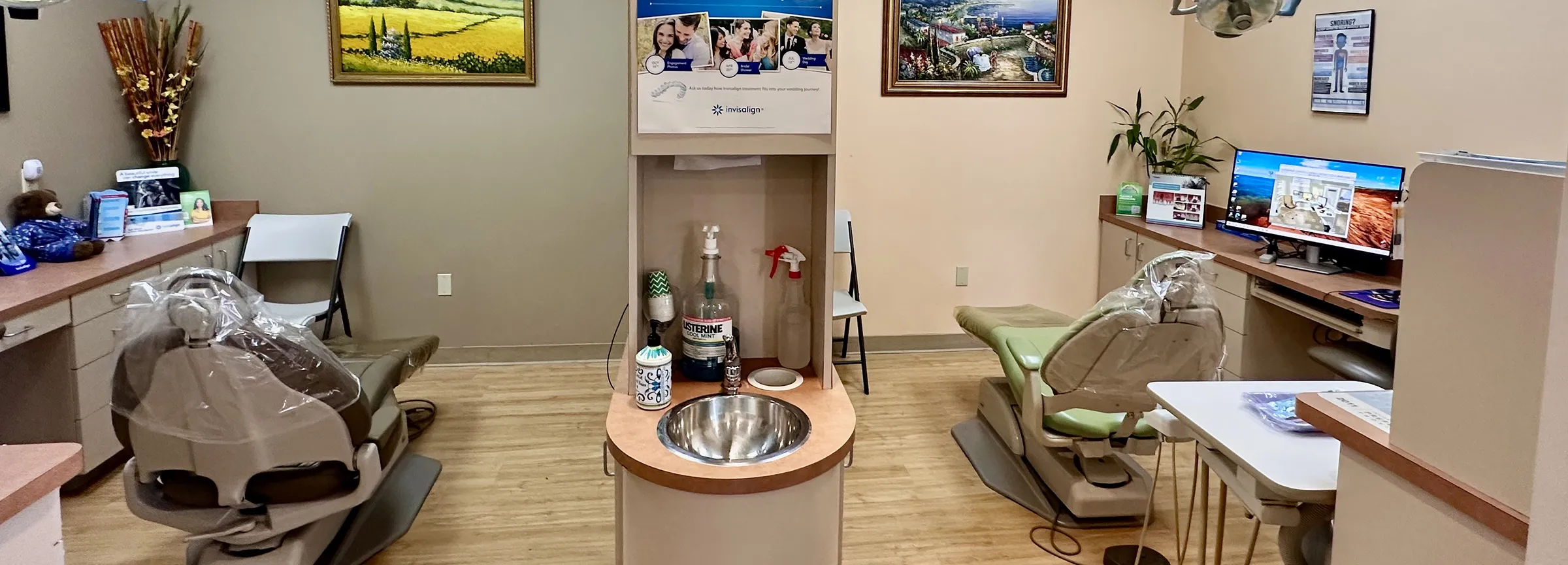 Dental office with two patient chairs, a central sink station, computer workstation, and framed artwork on beige walls.