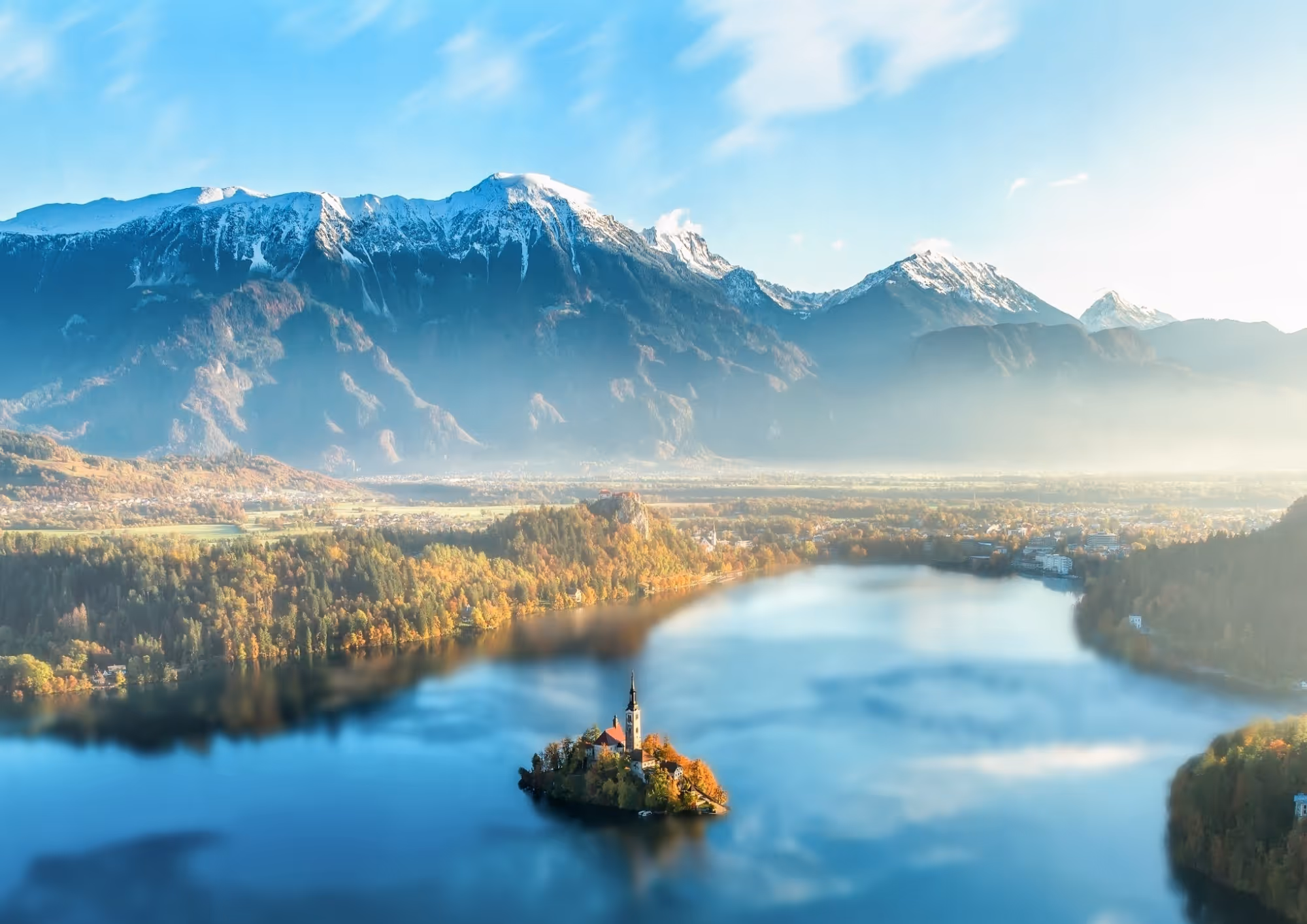 A serene landscape featuring a lake with an island and a church tower, surrounded by colorful autumn foliage and snow-capped mountains under a clear blue sky.