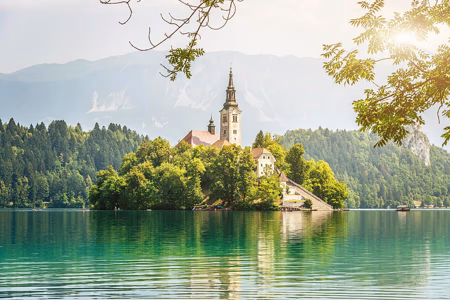 A serene landscape featuring a lake with an island and a church tower, surrounded by colorful autumn foliage and snow-capped mountains under a clear blue sky.