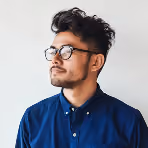 Young man with black curly hair, glasses, and a blue button-up shirt looking to the side against a light background.