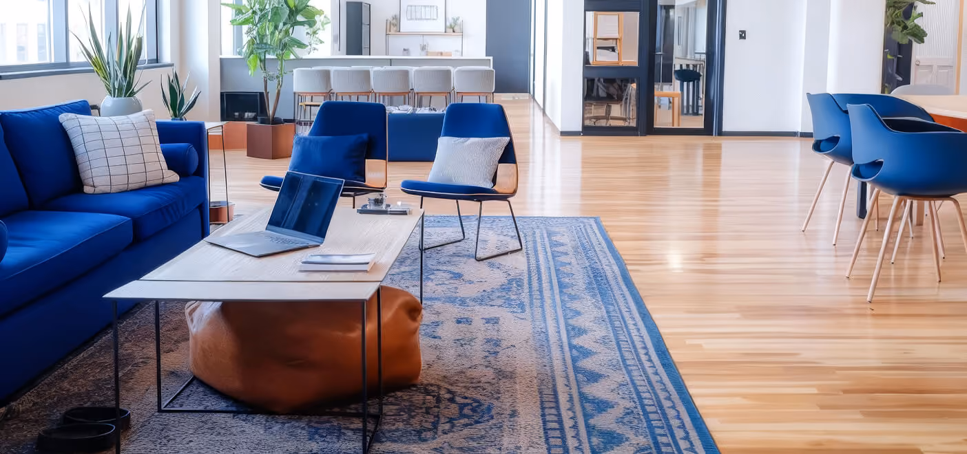 Modern office lounge with blue sofa, two blue chairs with pillows, wooden table with laptop, and patterned blue rug on light wood floor.