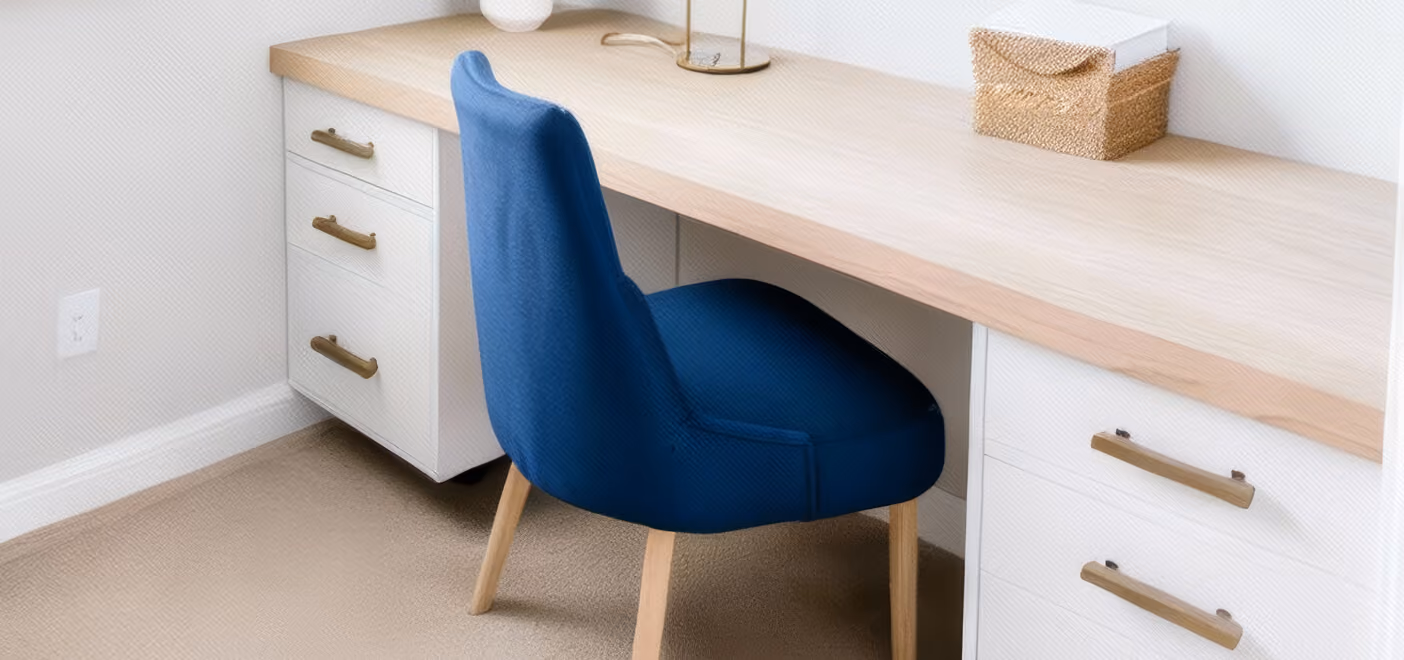 Blue upholstered chair with wooden legs tucked under a light wood desk with white drawers and brass handles.