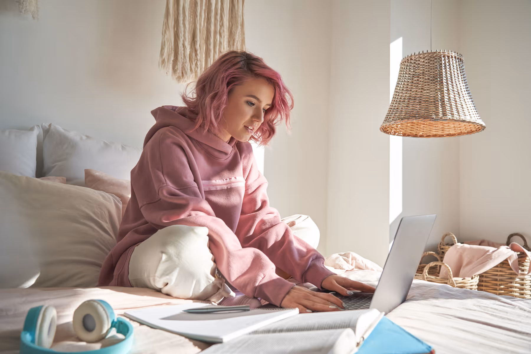 Teenage girl with pink hair sitting on her bed interacting with her laptop.