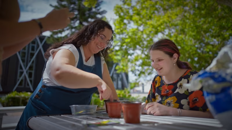 Two young women smiling and planting seeds in pots outdoors at a round table on a sunny day.