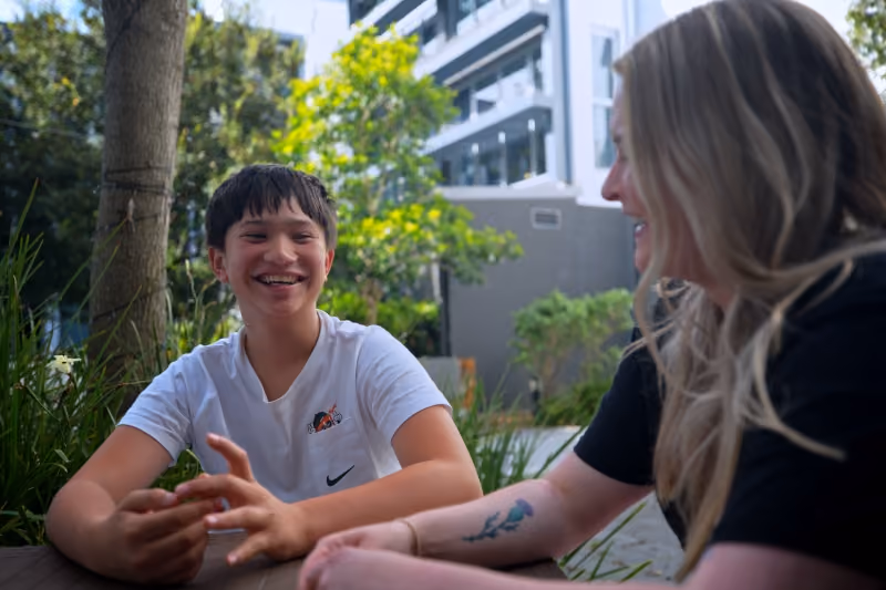 Teen boy smiling and talking with a woman, outdoors with greenery and buildings behind.