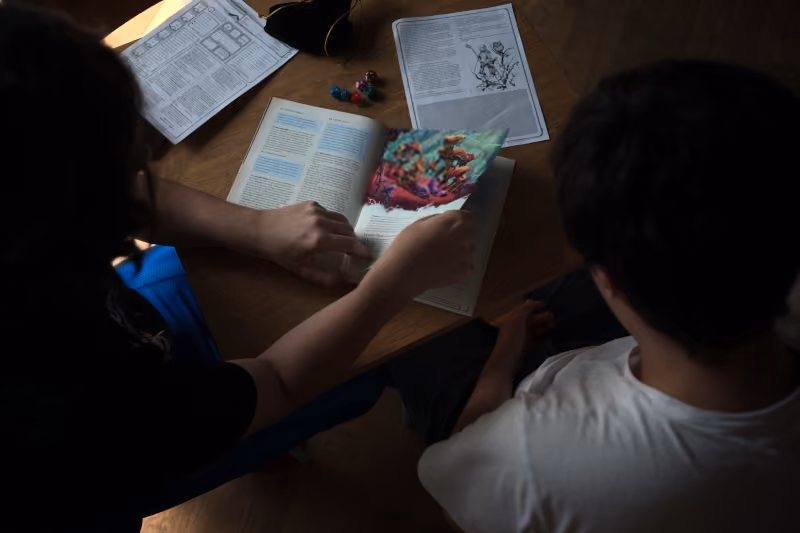 Two people sitting at a table looking at a game book with papers and colorful game pieces.