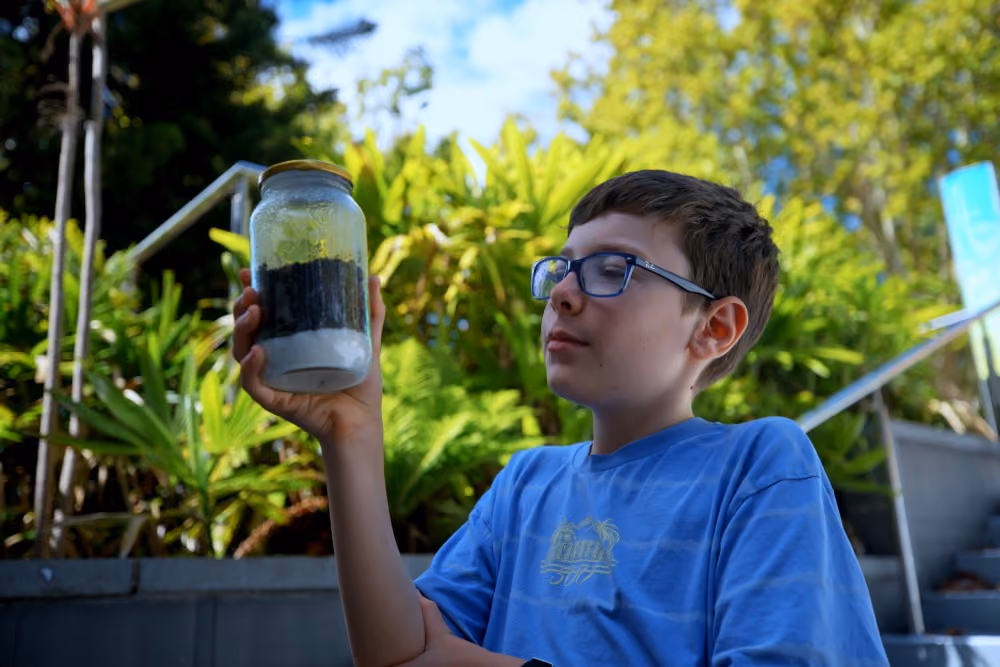 Boy wearing glasses holds a jar with soil layers while sitting outdoors with greenery behind him.