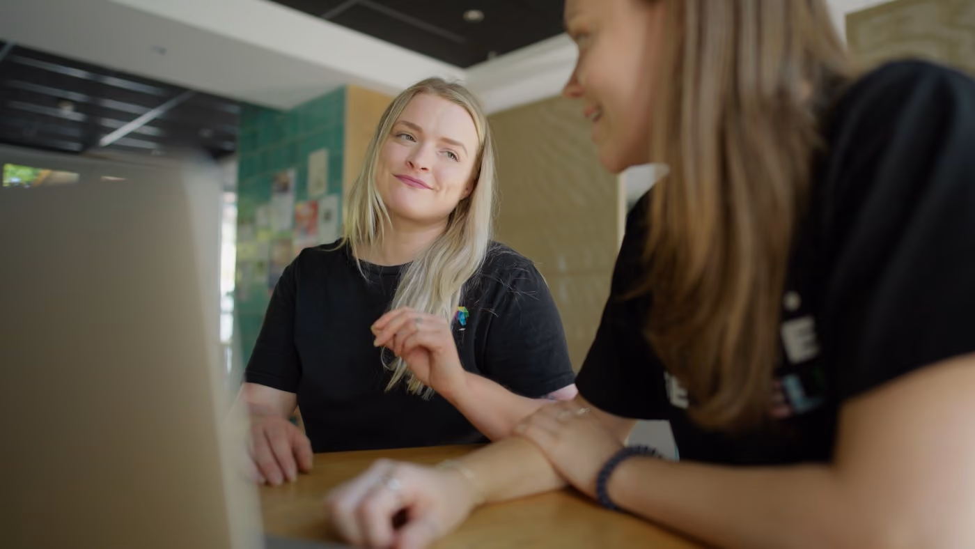 Two women smiling and talking at a table with a laptop in a casual indoor setting.