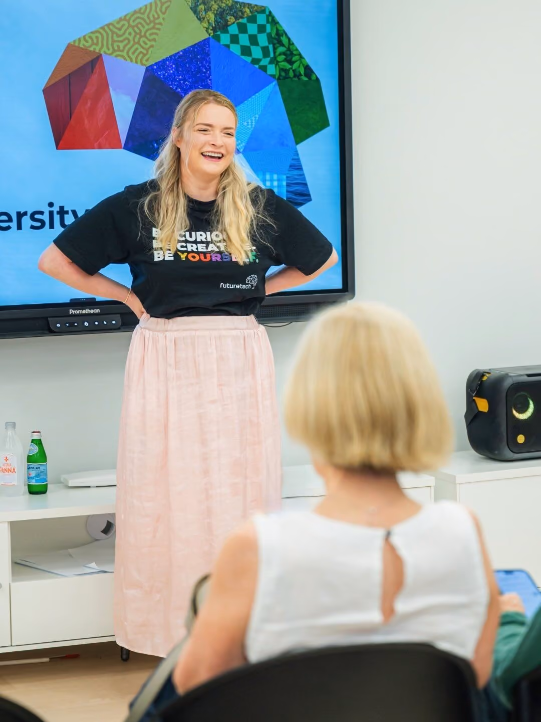Smiling woman in black shirt and pink skirt speaking in front of a screen with FutureTech logo.