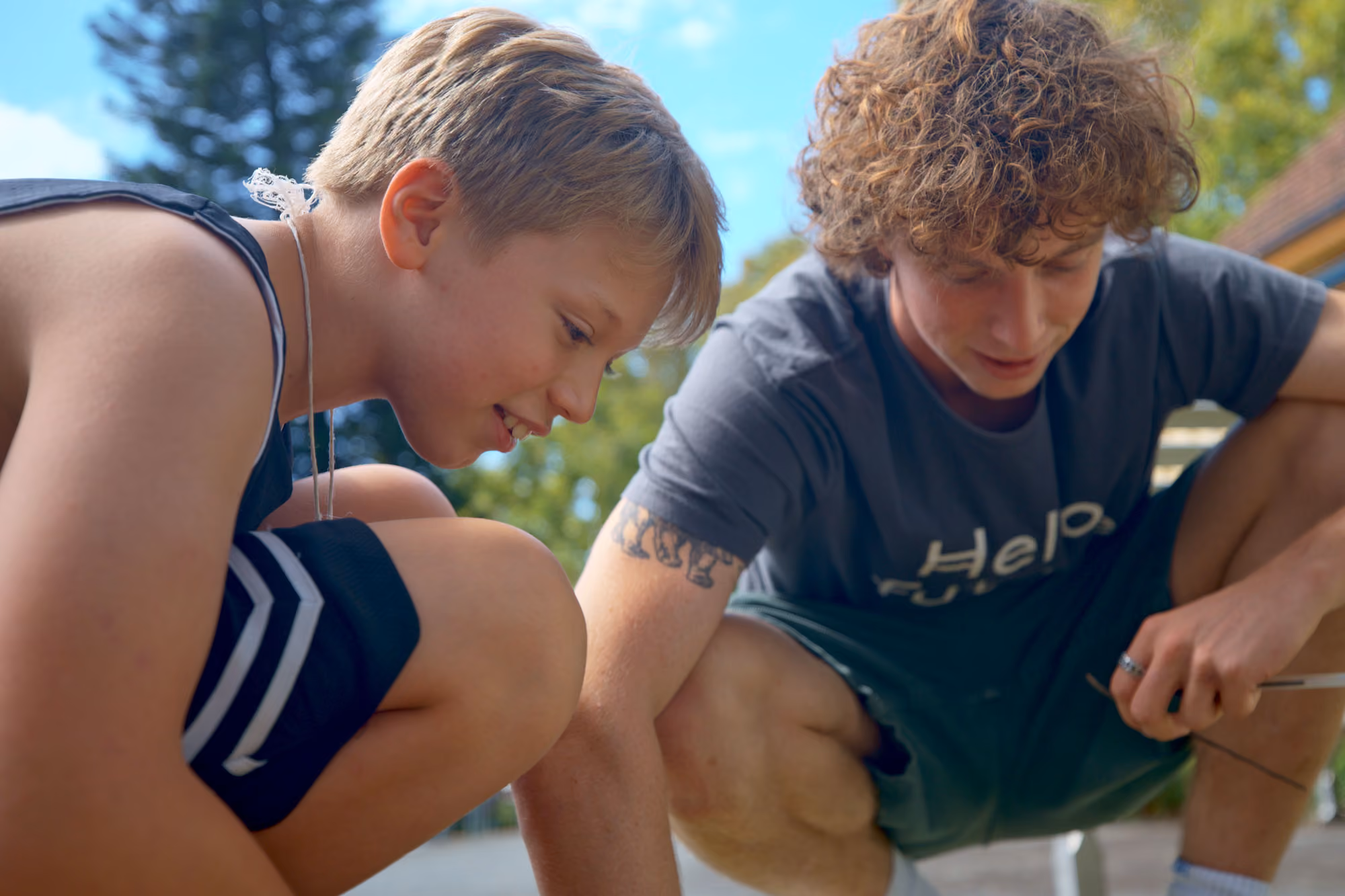 Two boys crouched outdoors, focused on an activity, one with blond hair and the other with curly hair.