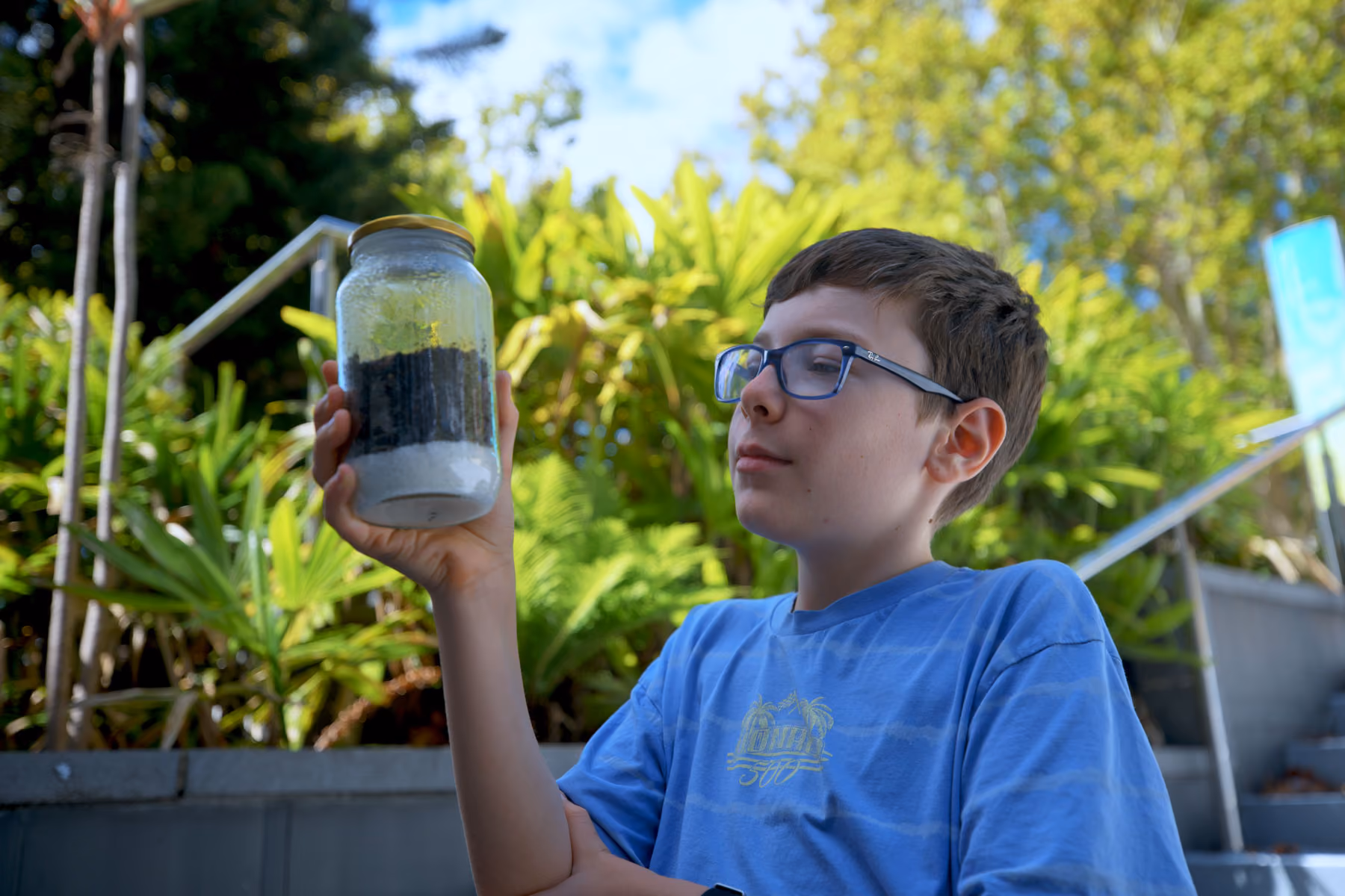 Boy wearing glasses holds a jar with soil layers while sitting outdoors with greenery behind him.