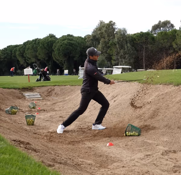 Edward Holland golfer hitting a shot from a sand bunker on a golf course with baskets of golf balls nearby.