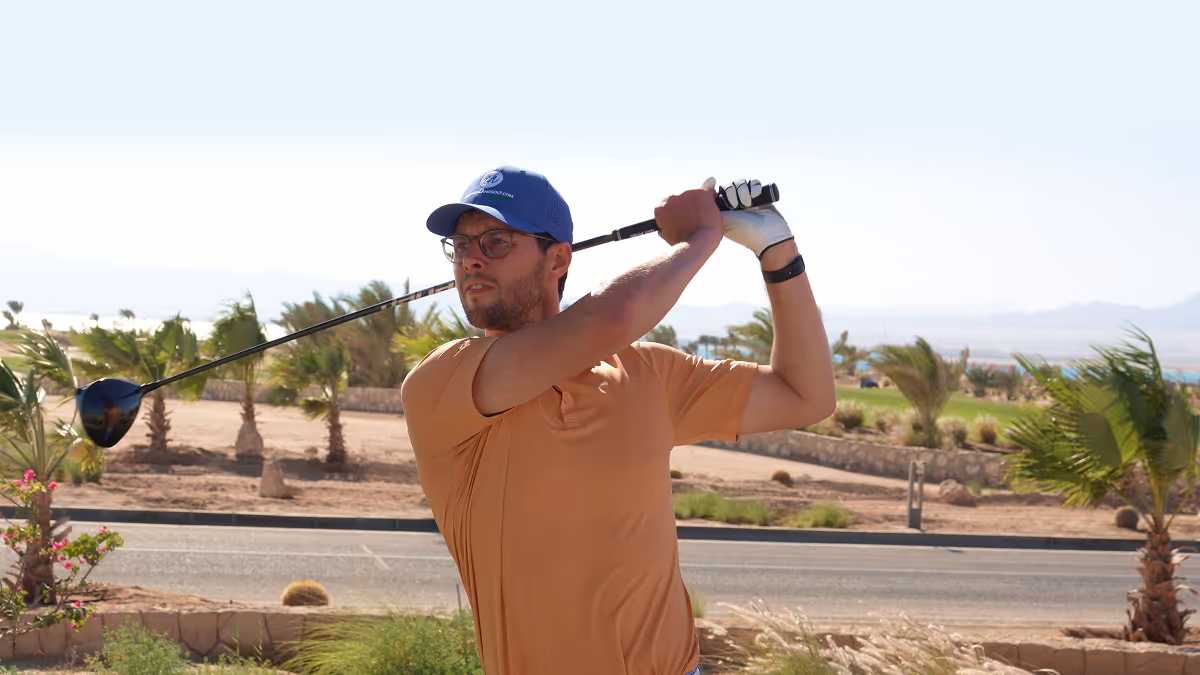 Edward Holland wearing glasses and blue cap swinging a golf club outdoors with palm trees and a road in the background.