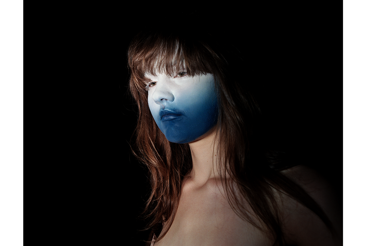 Studio portrait of a model styled by Bianca Fleisch. Model has a blue face, lights are focusing the face. Surrounded by darkness.