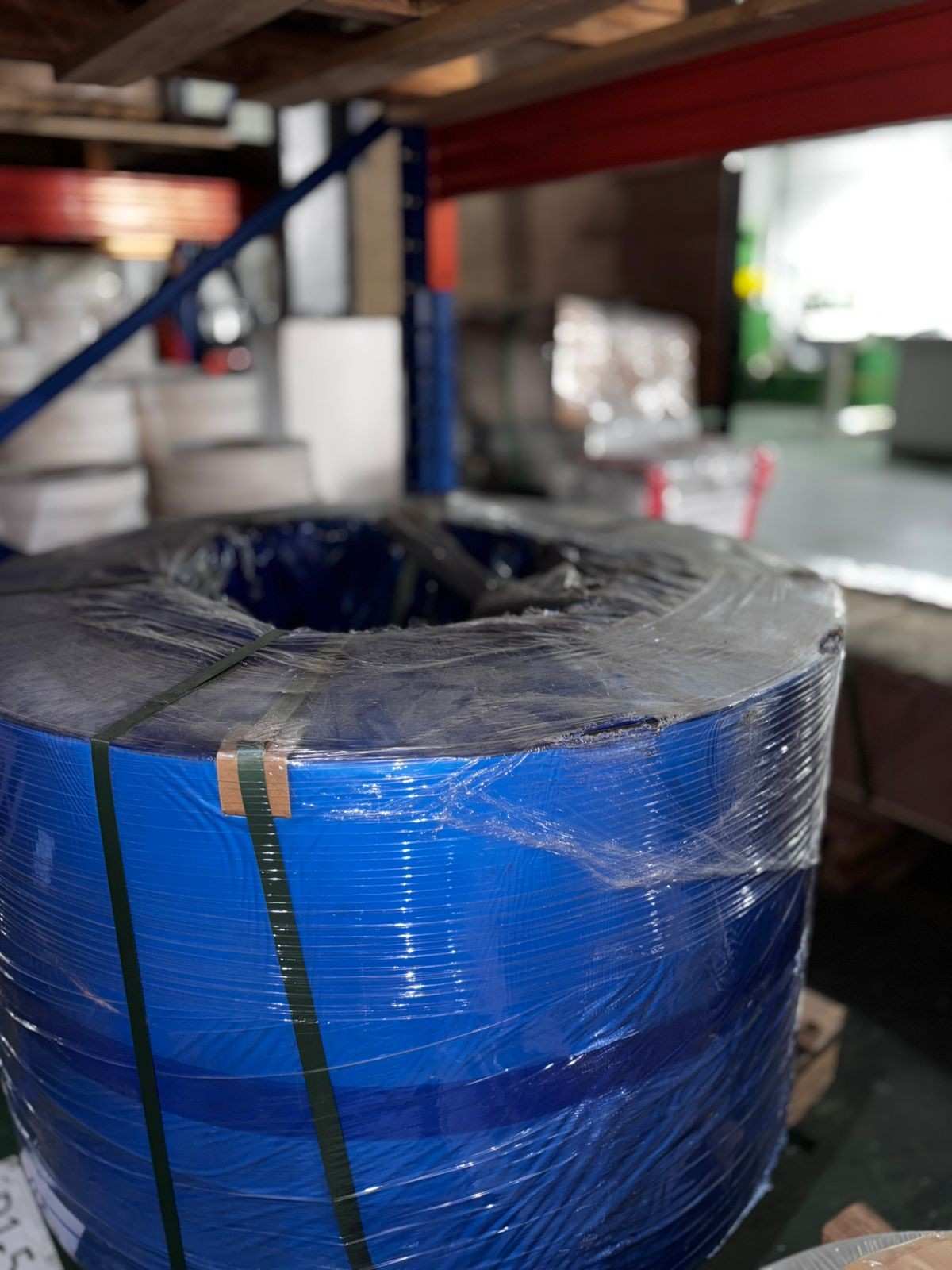 Large coil of blue industrial material wrapped in plastic and secured with black strapping in a warehouse setting.