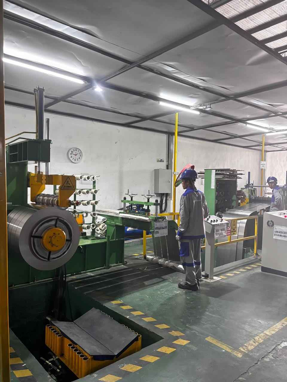 Industrial setting with two workers in safety helmets and gray-blue uniforms operating machinery next to metal coil and conveyor system.