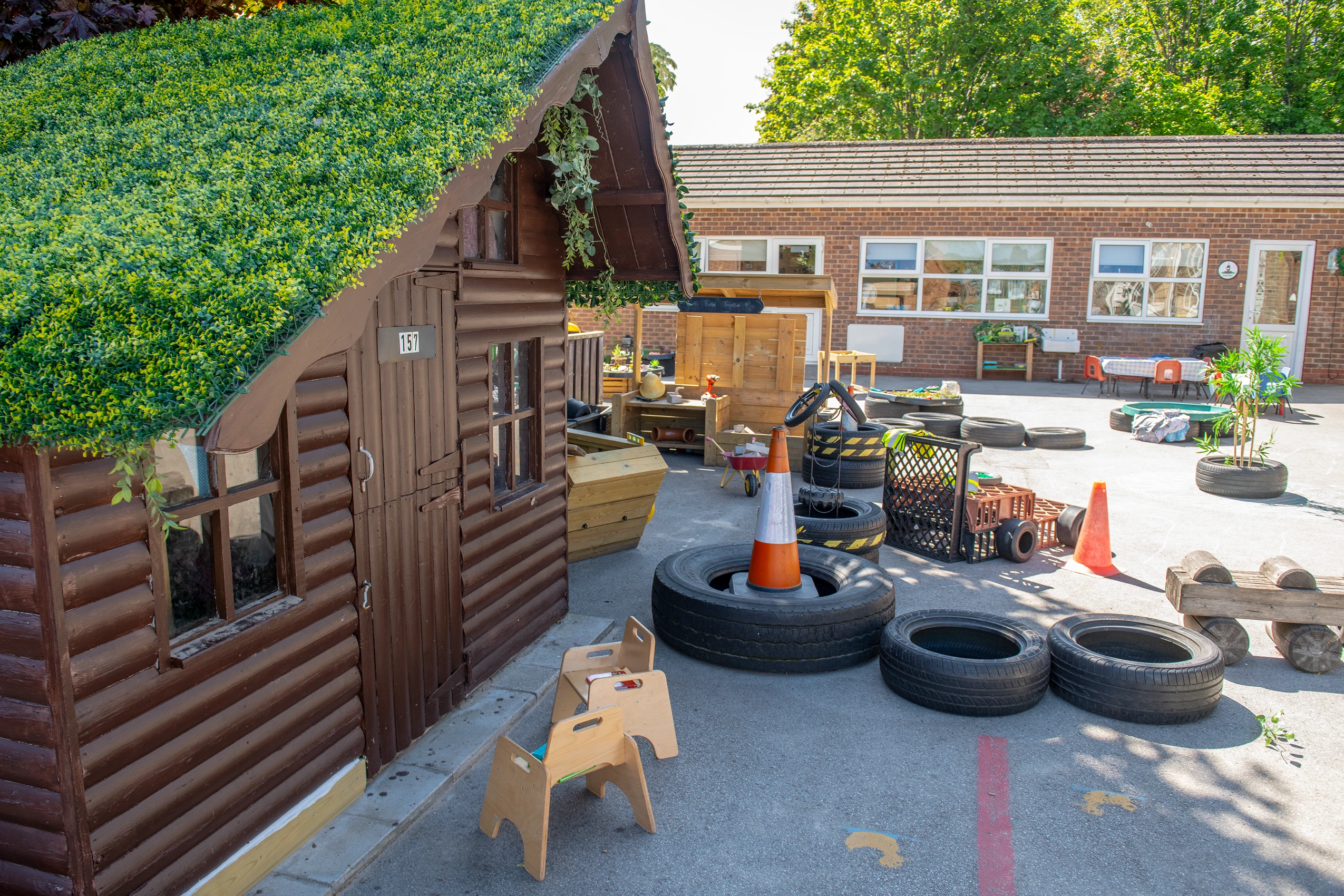 A small wooden playhouse with a green foliage roof, dark brown log walls, and a sign with the number '157'.