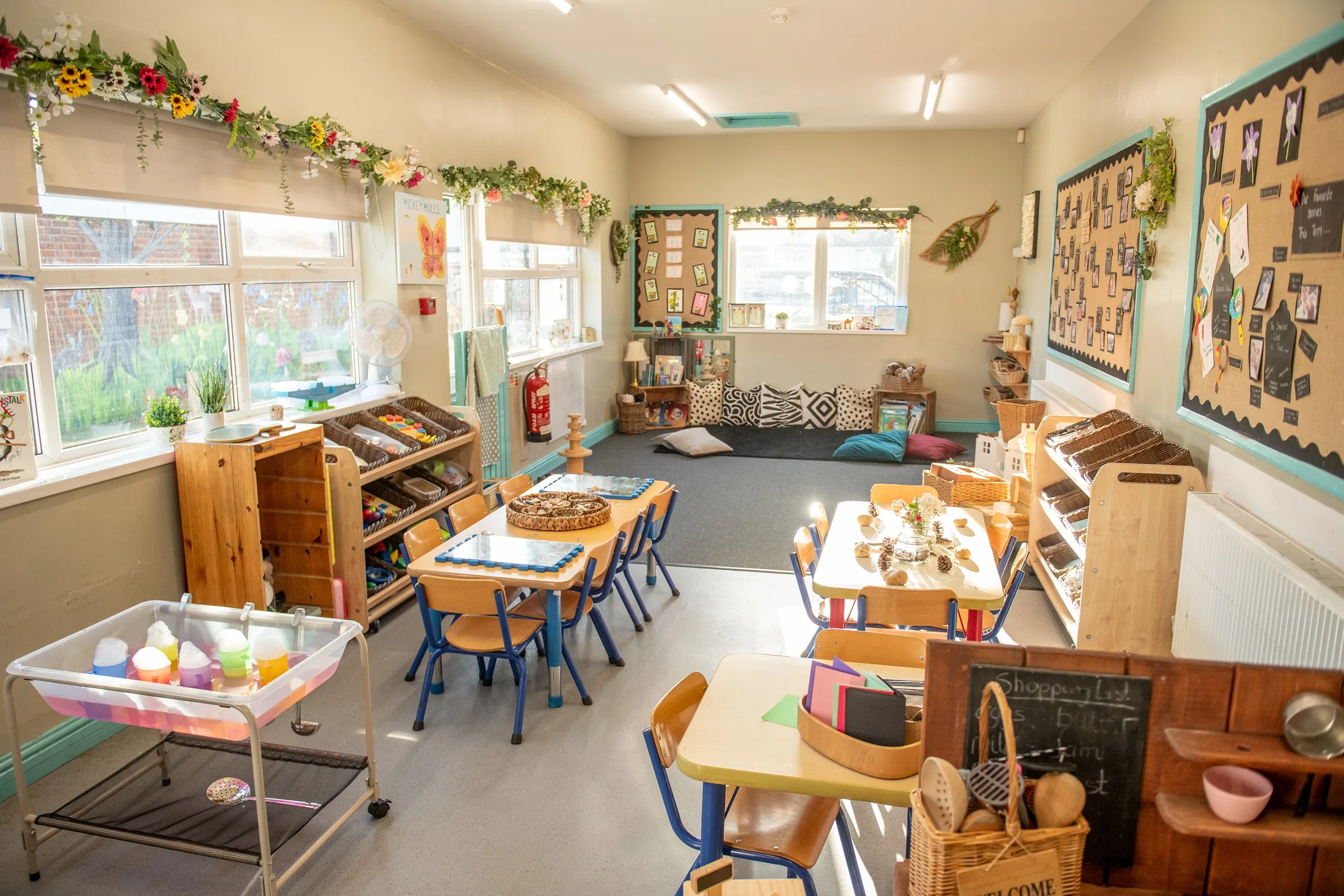 A brightly lit classroom with small tables, chairs, shelves of toys, and colorful artwork on the walls.