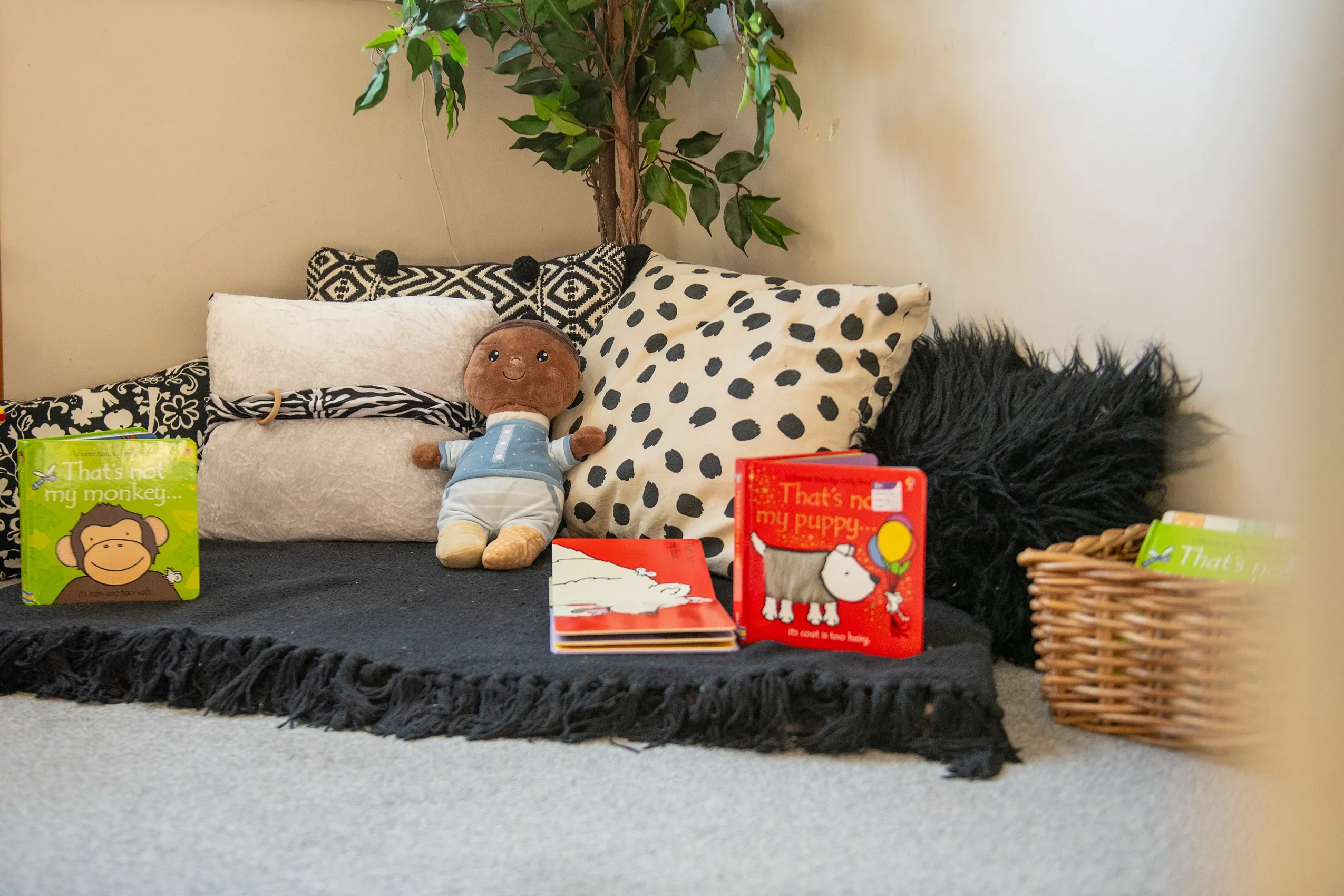 A brown-skinned baby doll in a light blue shirt and striped pants, sitting on a black fringed blanket surrounded by pillows and children's books.