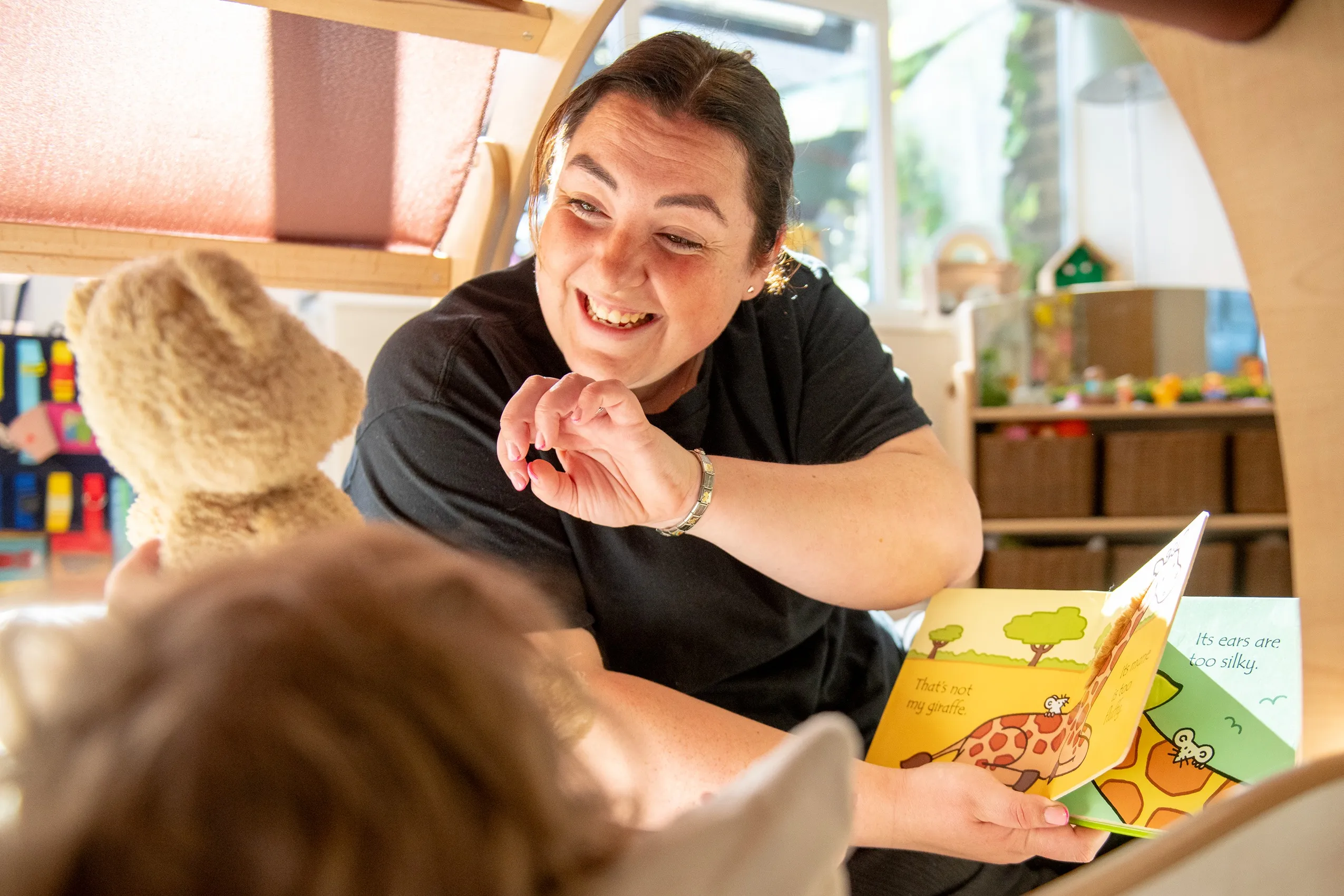 A smiling woman reading a book with a giraffe illustration to a child lying down.