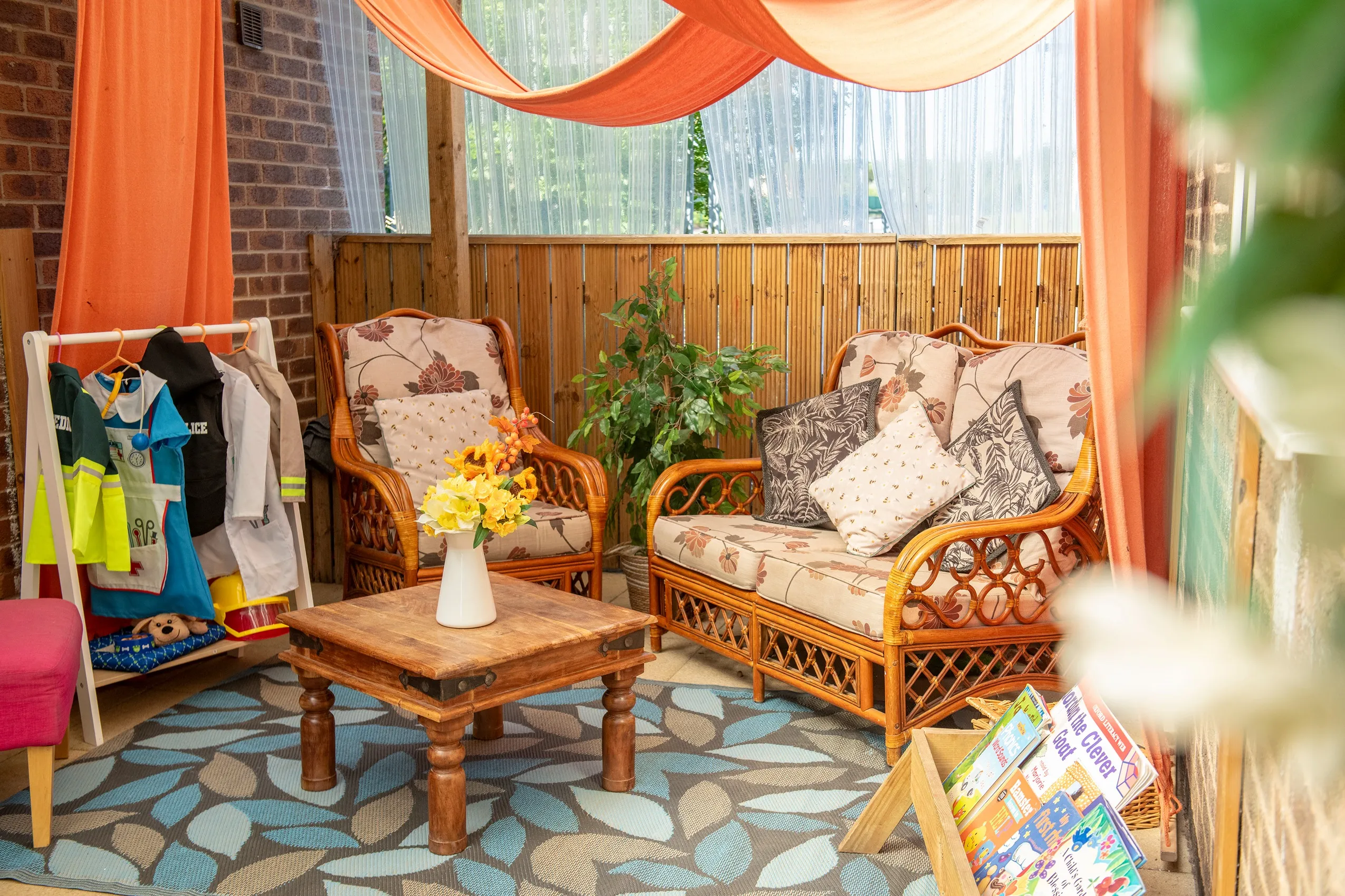 A cozy outdoor seating area with wicker chairs, a loveseat, floral cushions, a wooden coffee table with yellow flowers, and a leaf-patterned rug.