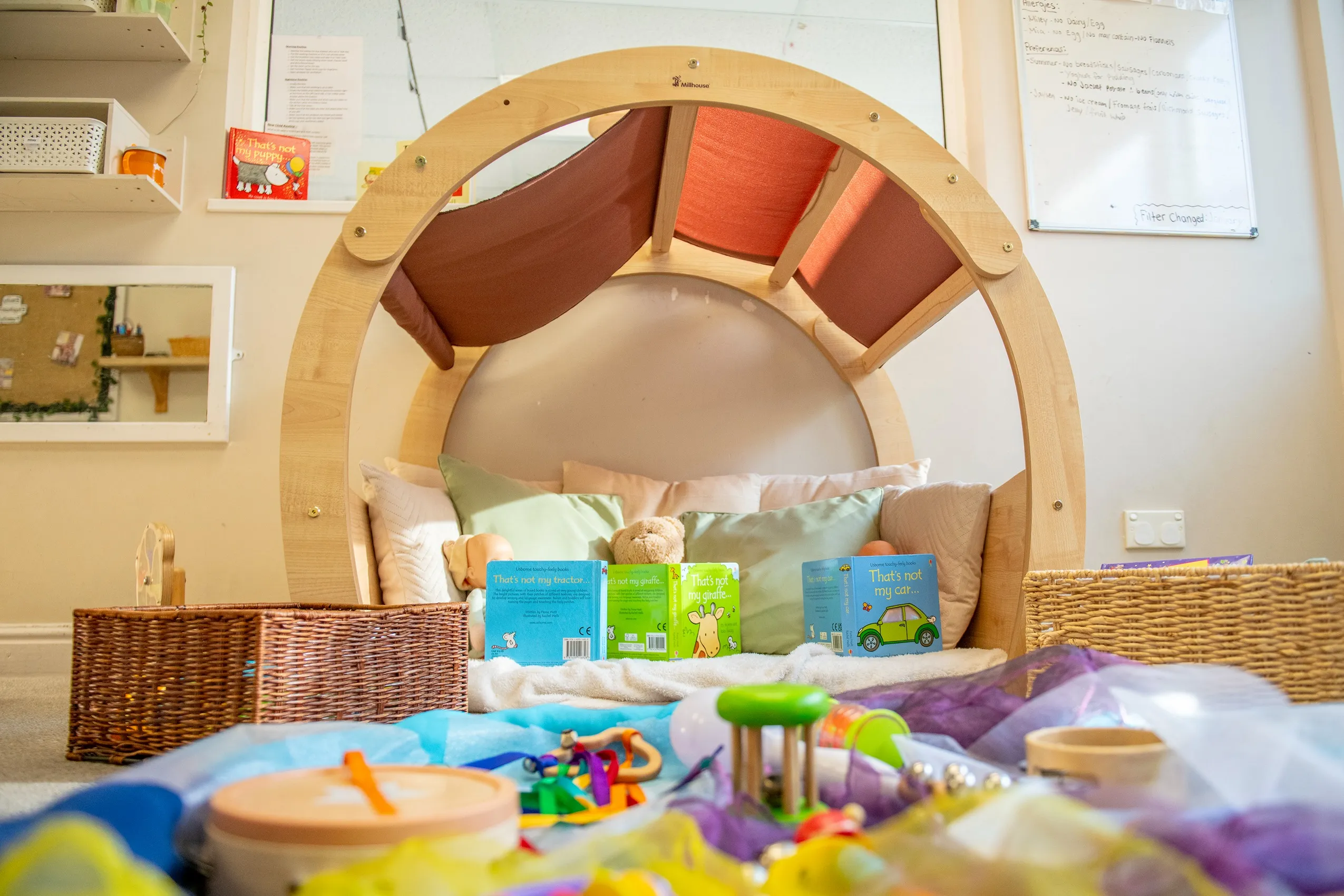 A wooden play structure with fabric panels, pillows, children's books, and various toys on a soft fabric, with woven baskets on the sides.