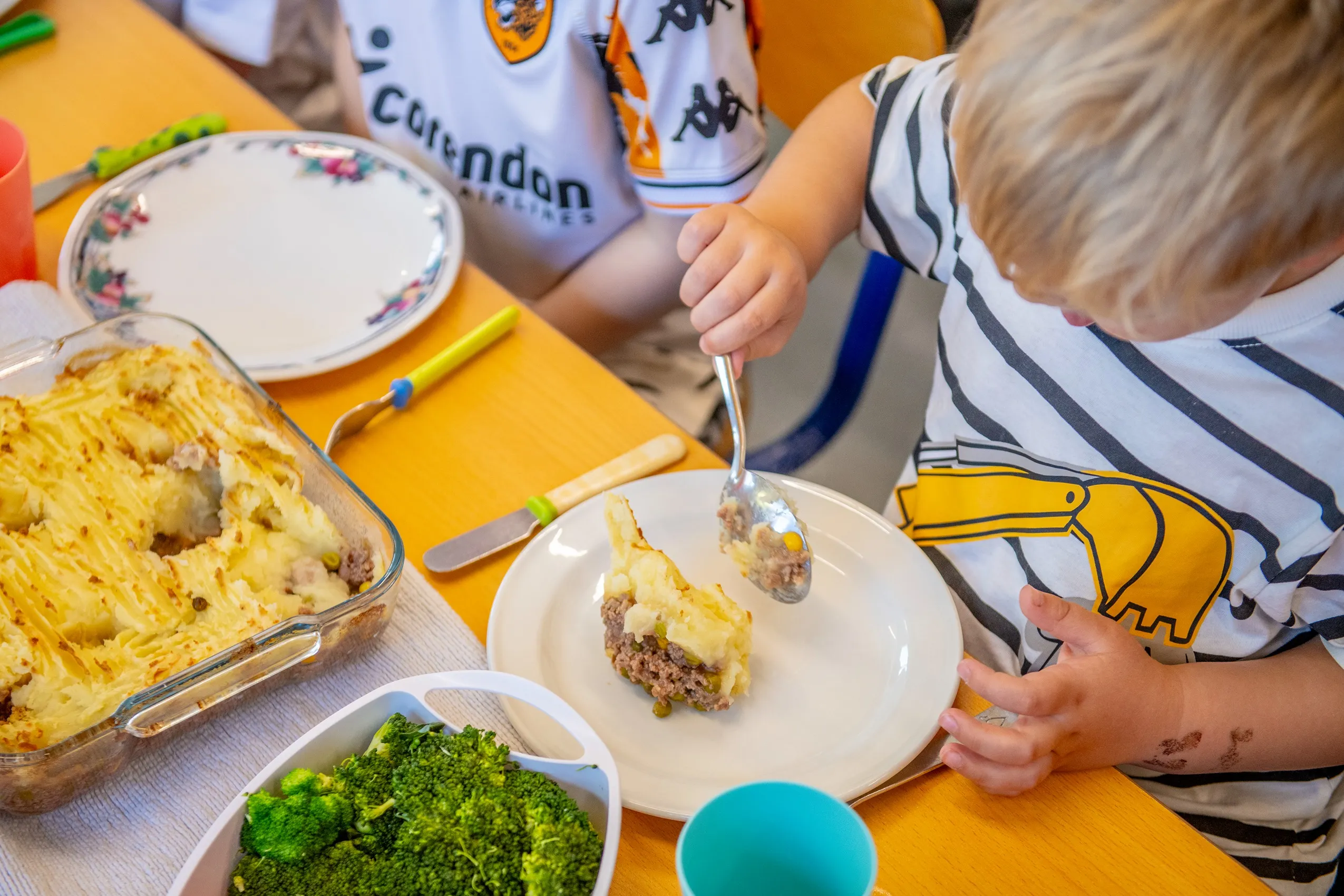 A child eating shepherd's pie from a white plate, with a baking dish of more pie and a bowl of broccoli nearby.