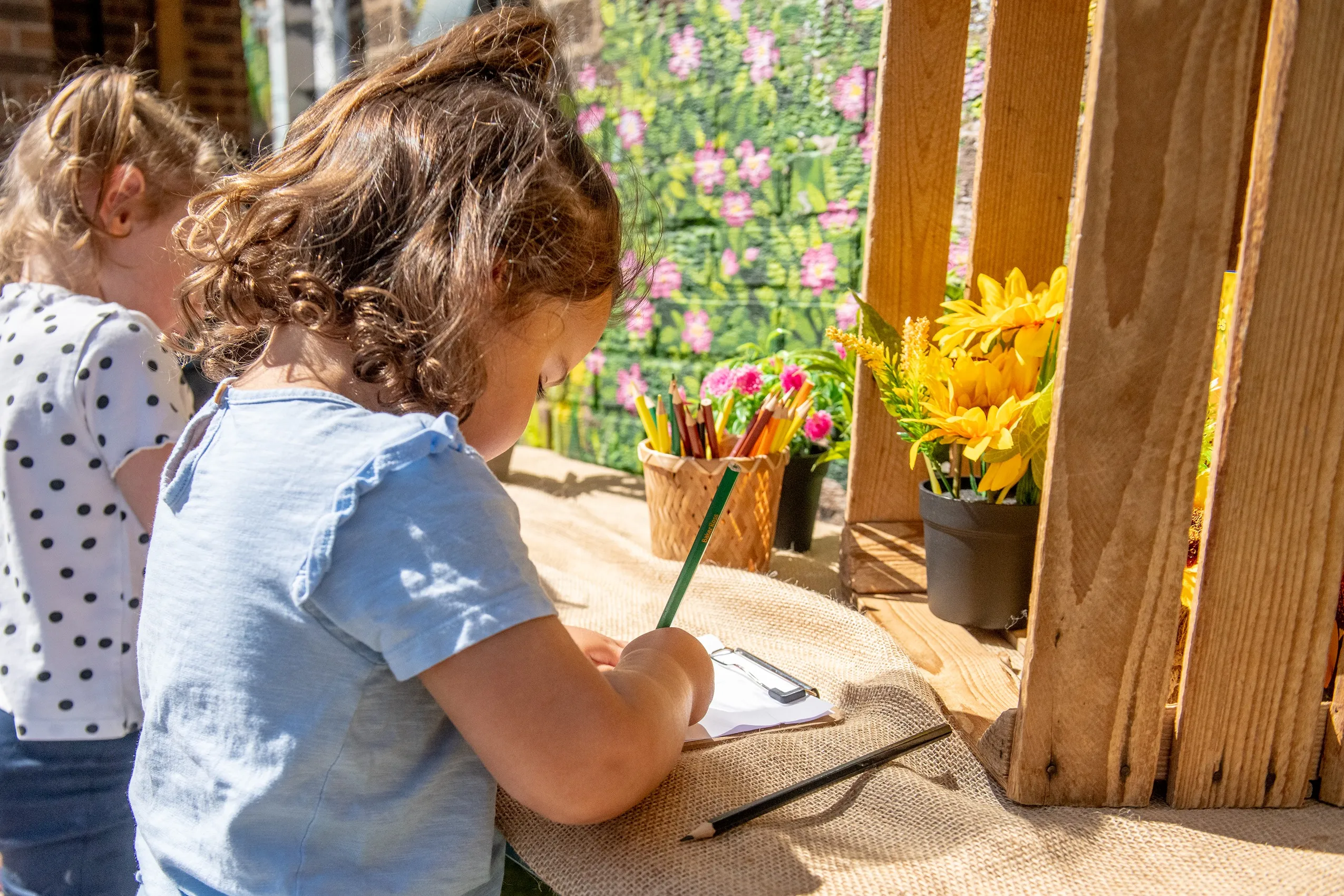 A young girl with curly brown hair drawing on a clipboard at a burlap-covered table, with another girl and flowers in the background.
