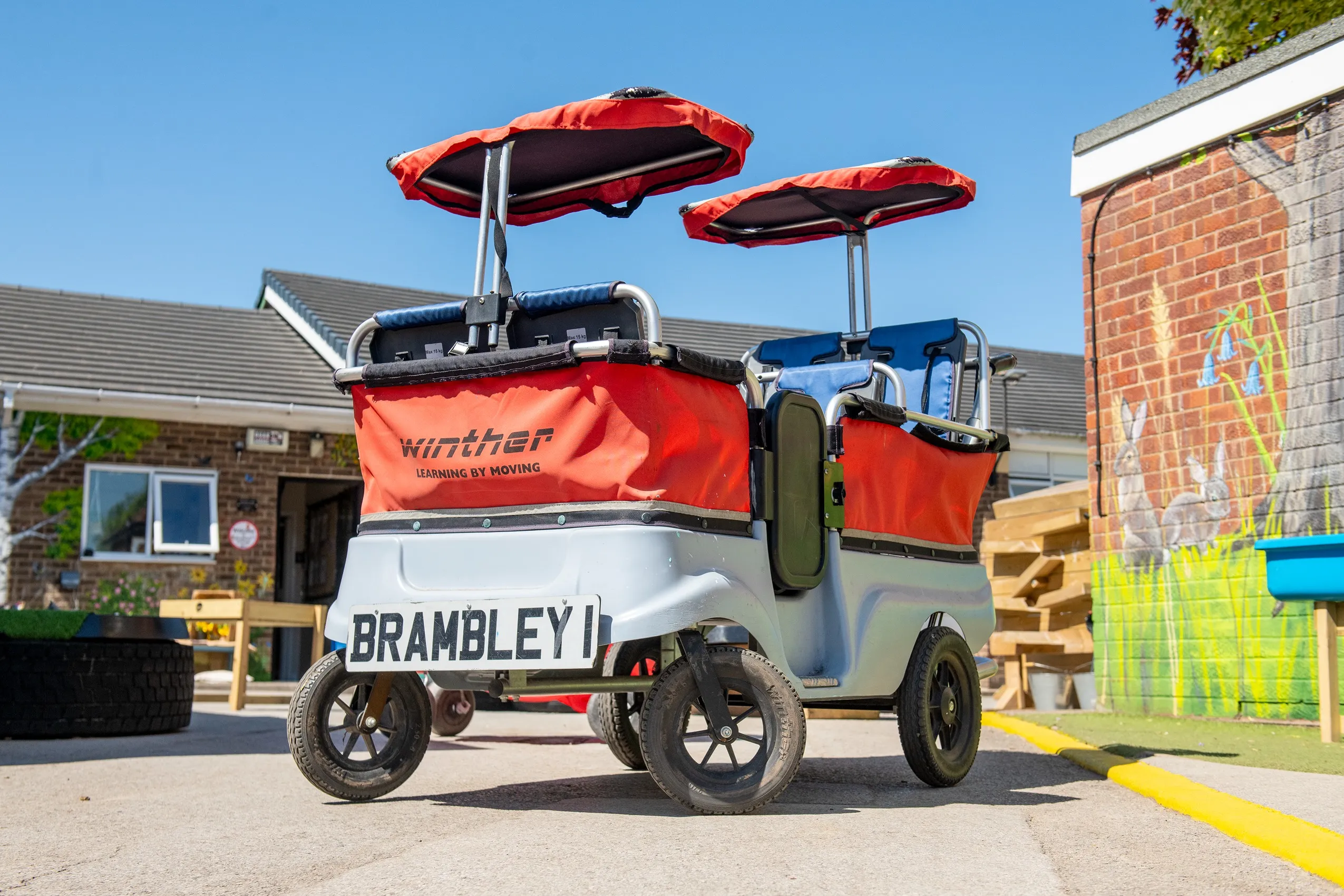 A four-wheeled children's cart with two seats, blue cushions, red sunshades, and 'Winther LEARNING BY MOVING' text.