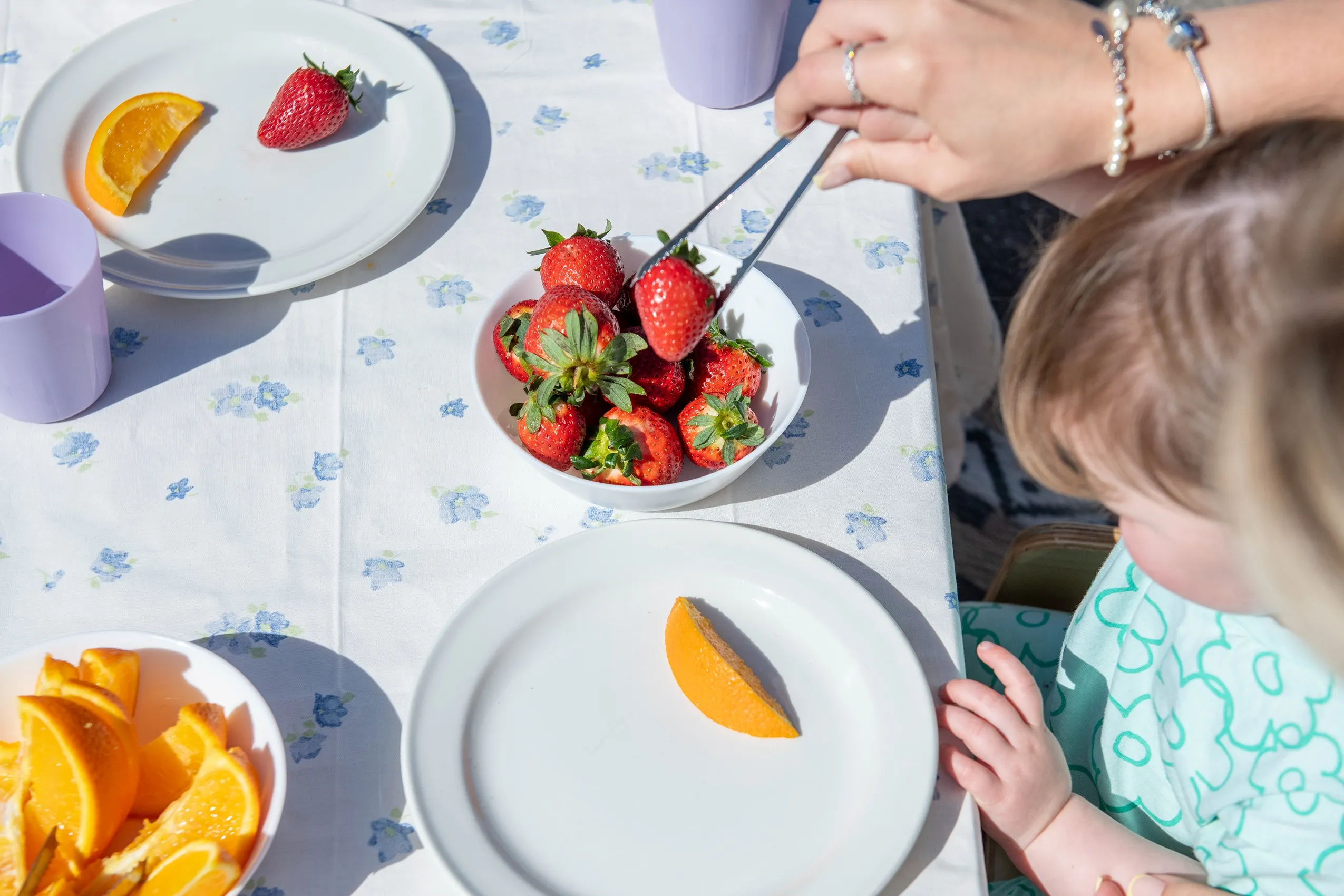 A white bowl filled with bright red strawberries, with tongs picking one up, on a blue floral tablecloth.