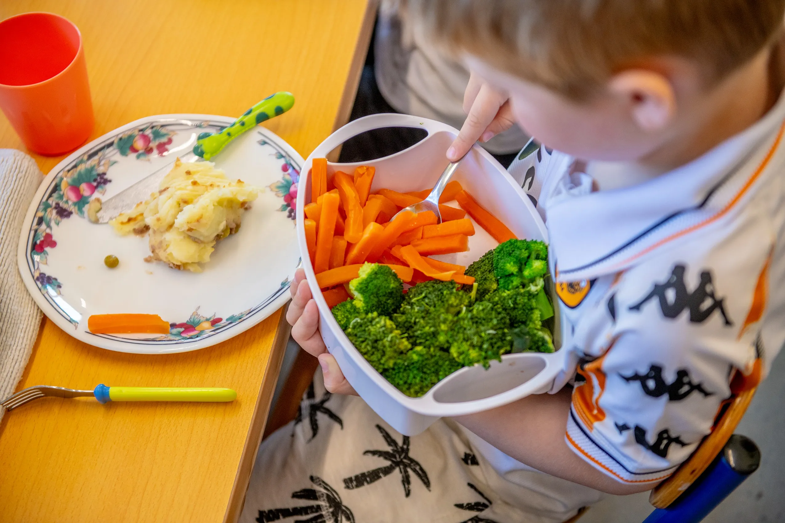 A child holding a white lunch box filled with orange carrot sticks and green broccoli florets.