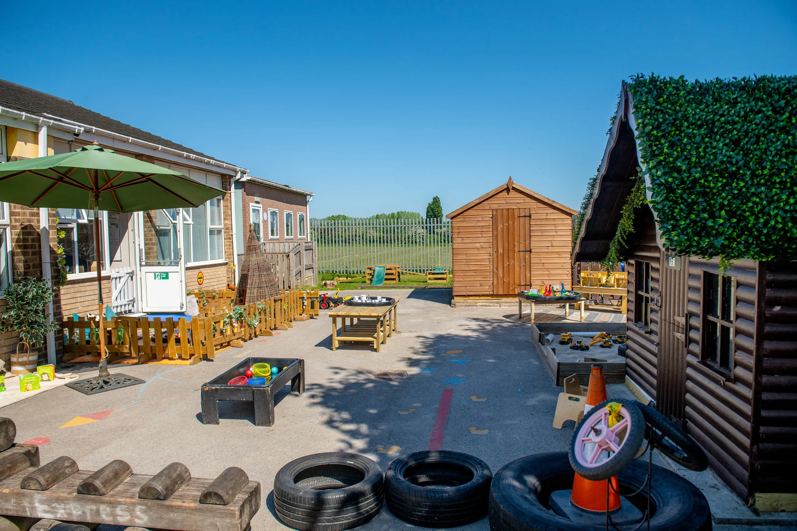 An outdoor play area with wooden sheds, tables, tires, sandboxes, and a playhouse, designed for imaginative play.