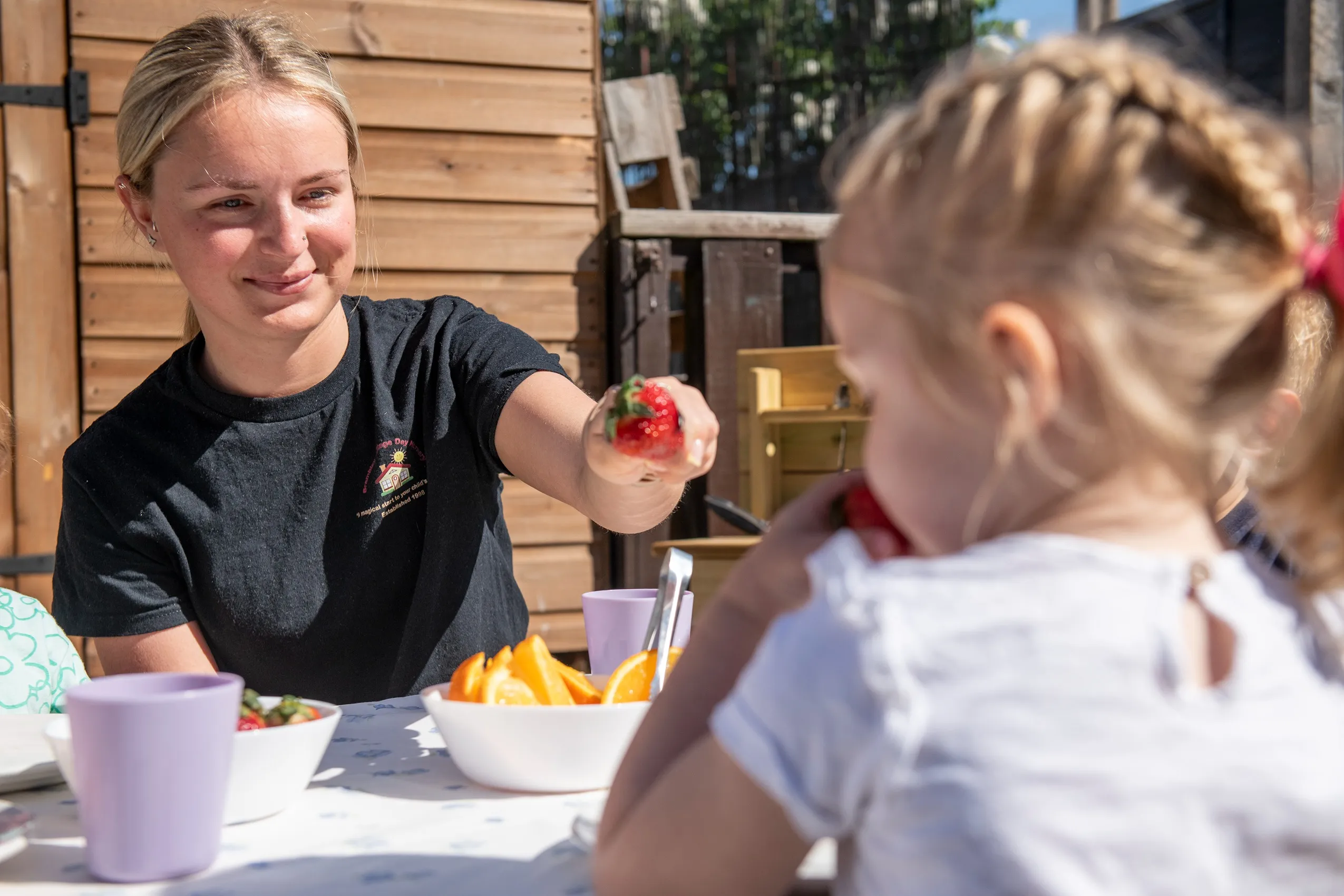 A woman holding out a bright red strawberry towards a child sitting at a table.