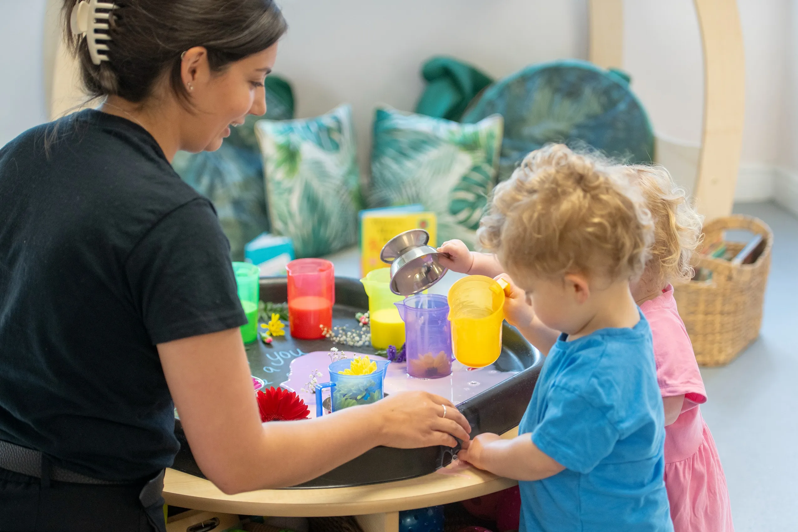 A woman and two children playing with colorful cups, pitchers, and flowers on a black tray on a wooden table.