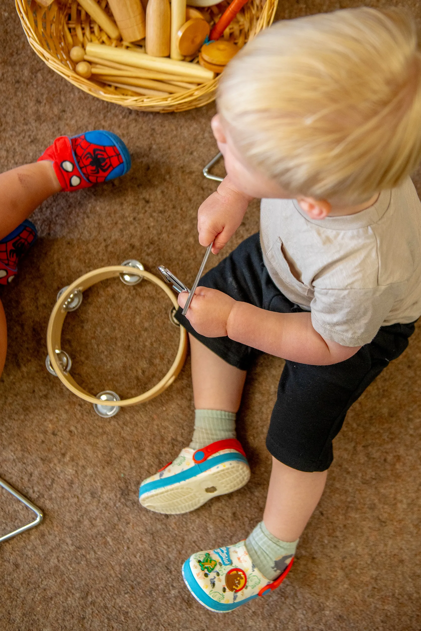 A wooden tambourine with metal jingles, lying on a brown carpet next to a child holding a metal stick.