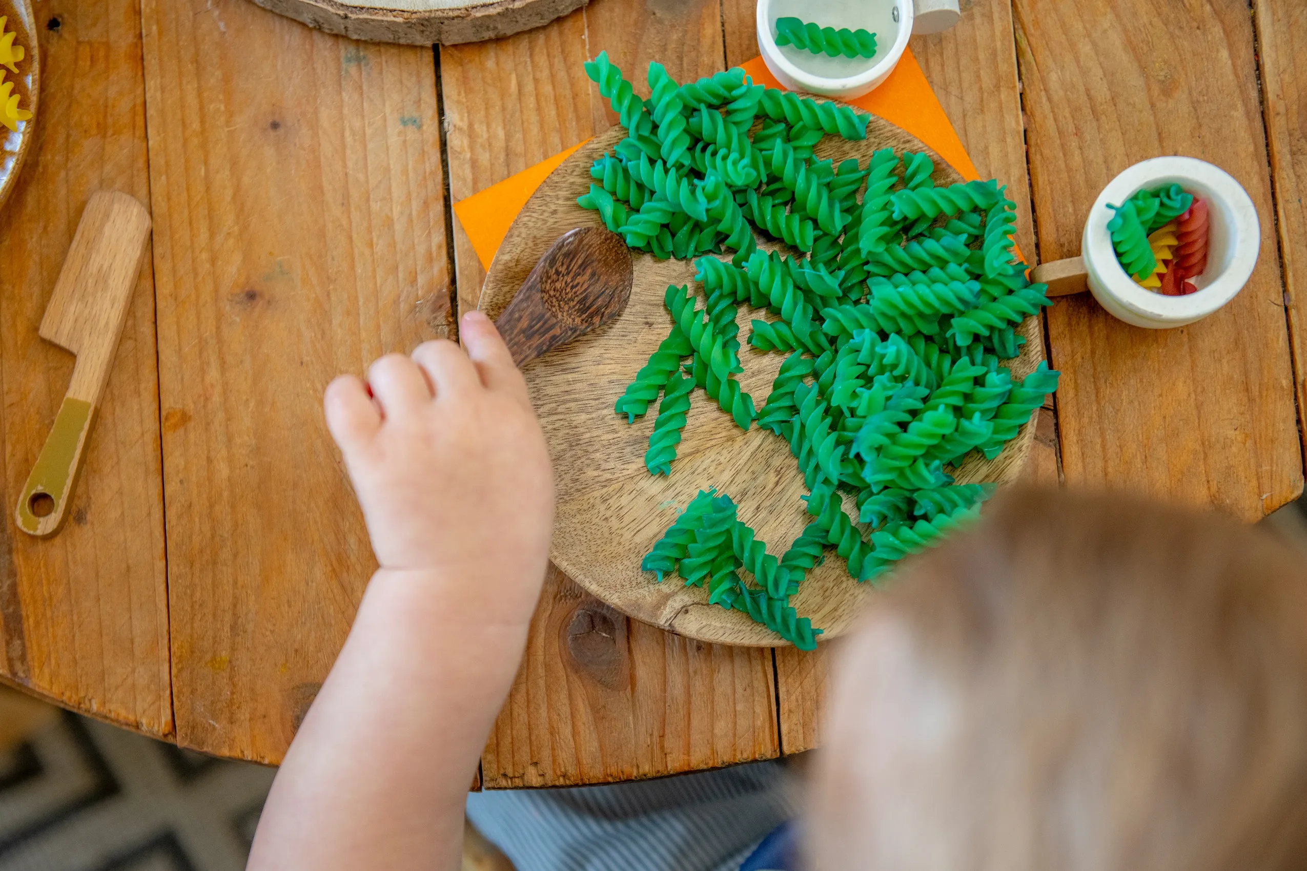 A pile of bright green spiral-shaped pasta with a wooden spoon on a round wooden plate.
