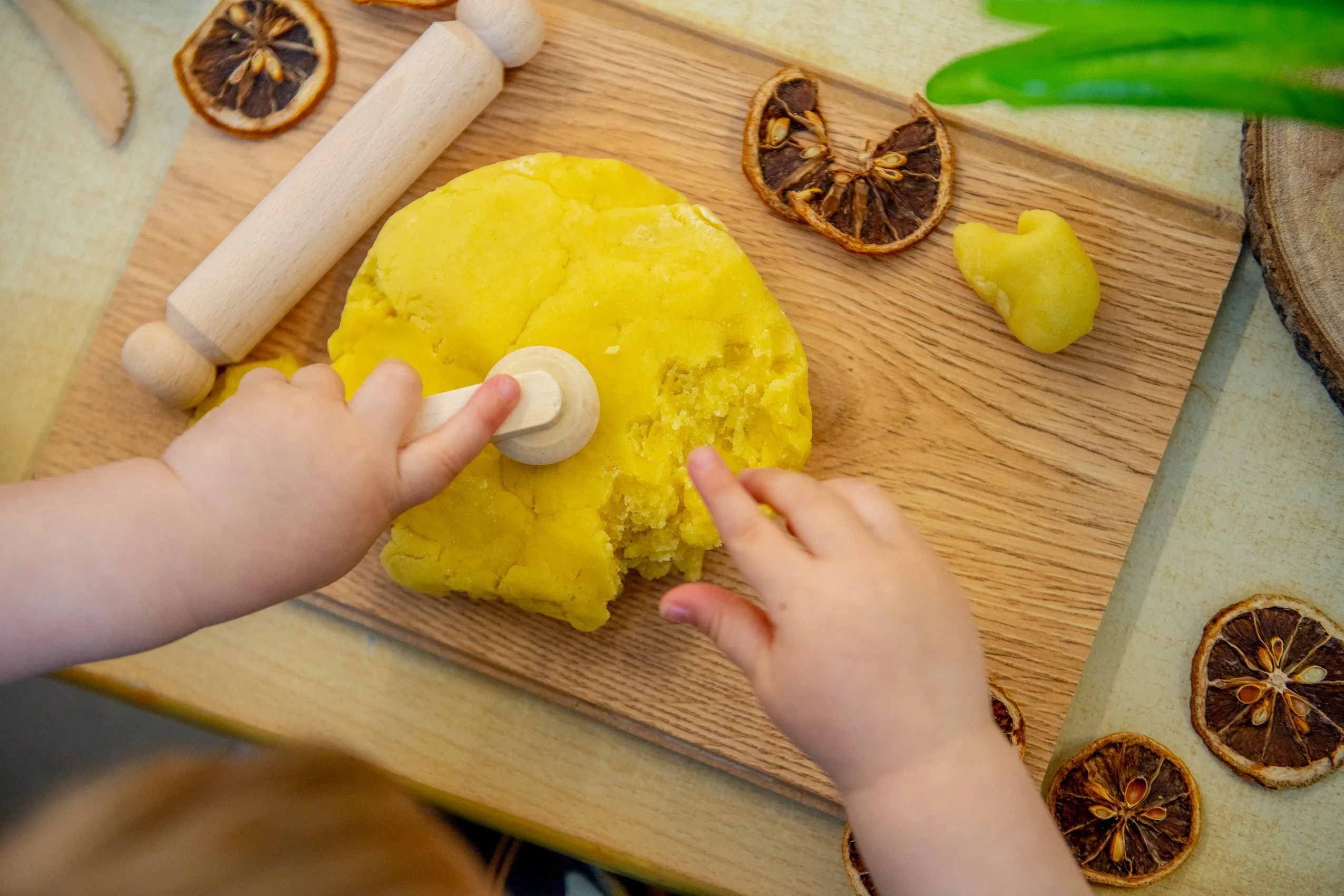 A child's hands interacting with bright yellow play dough on a wooden cutting board, surrounded by dried orange slices and a rolling pin.