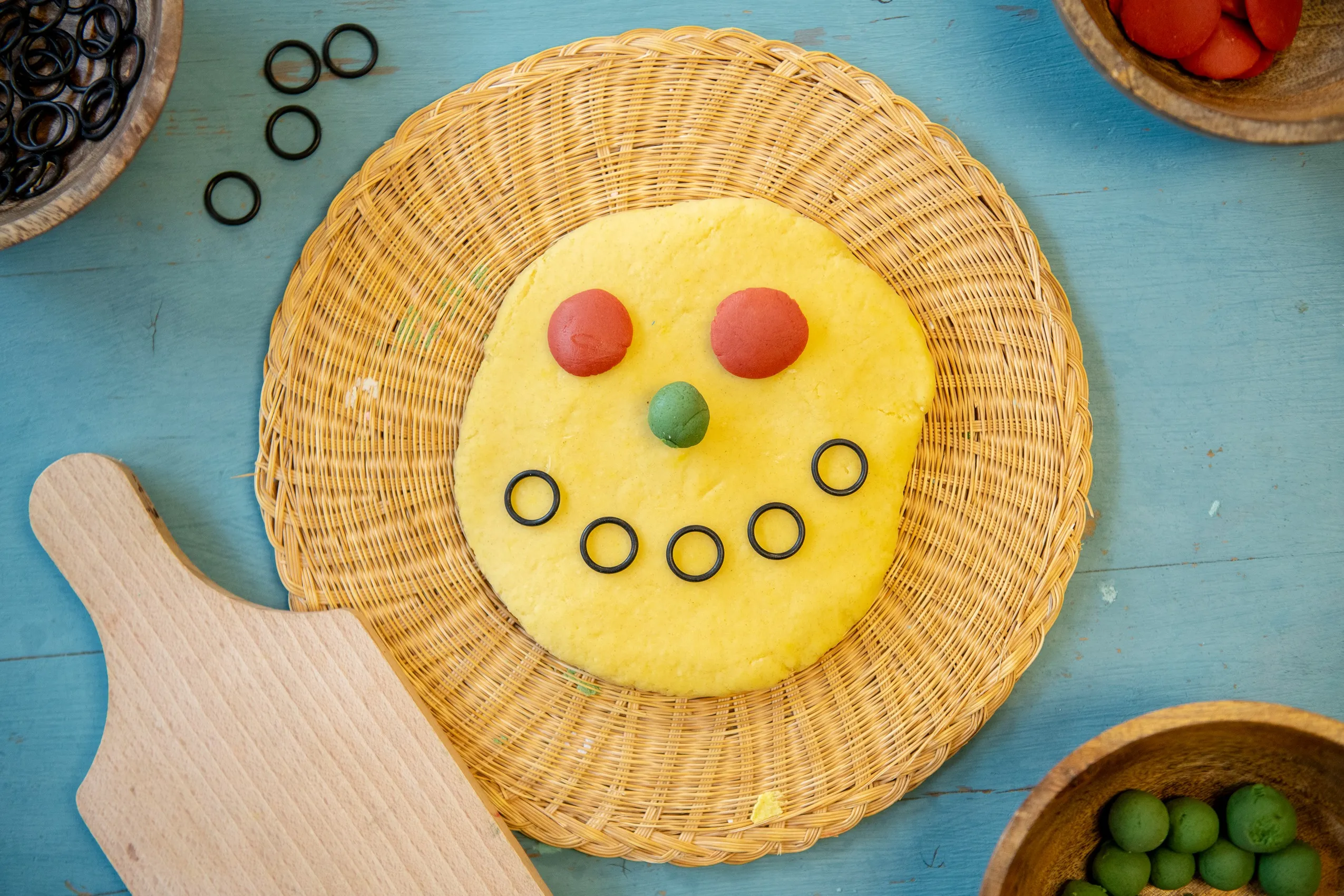 A smiley face made of yellow dough with red eyes, a green nose, and black rubber ring mouth on a woven placemat.