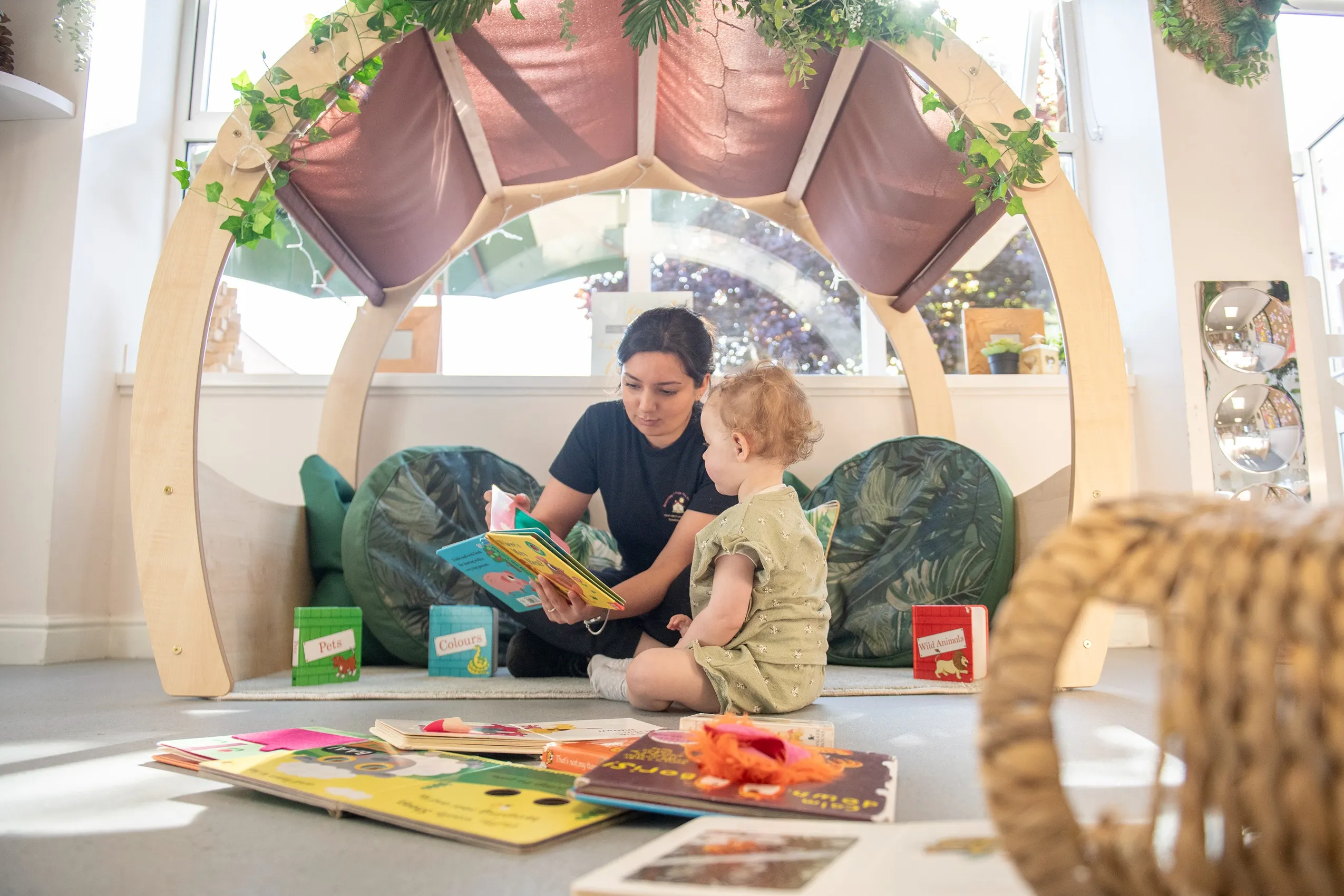 A woman reading a book to a child inside a wooden play structure with a fabric canopy and artificial greenery.