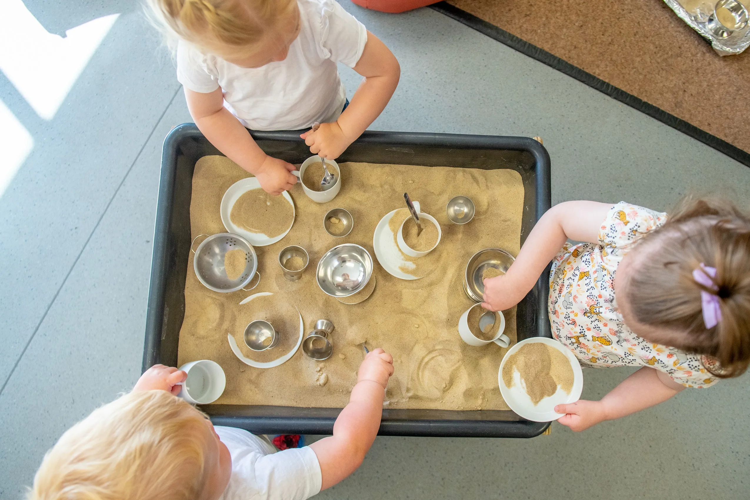 Three young children playing with sand and kitchen utensils in a black rectangular tray.