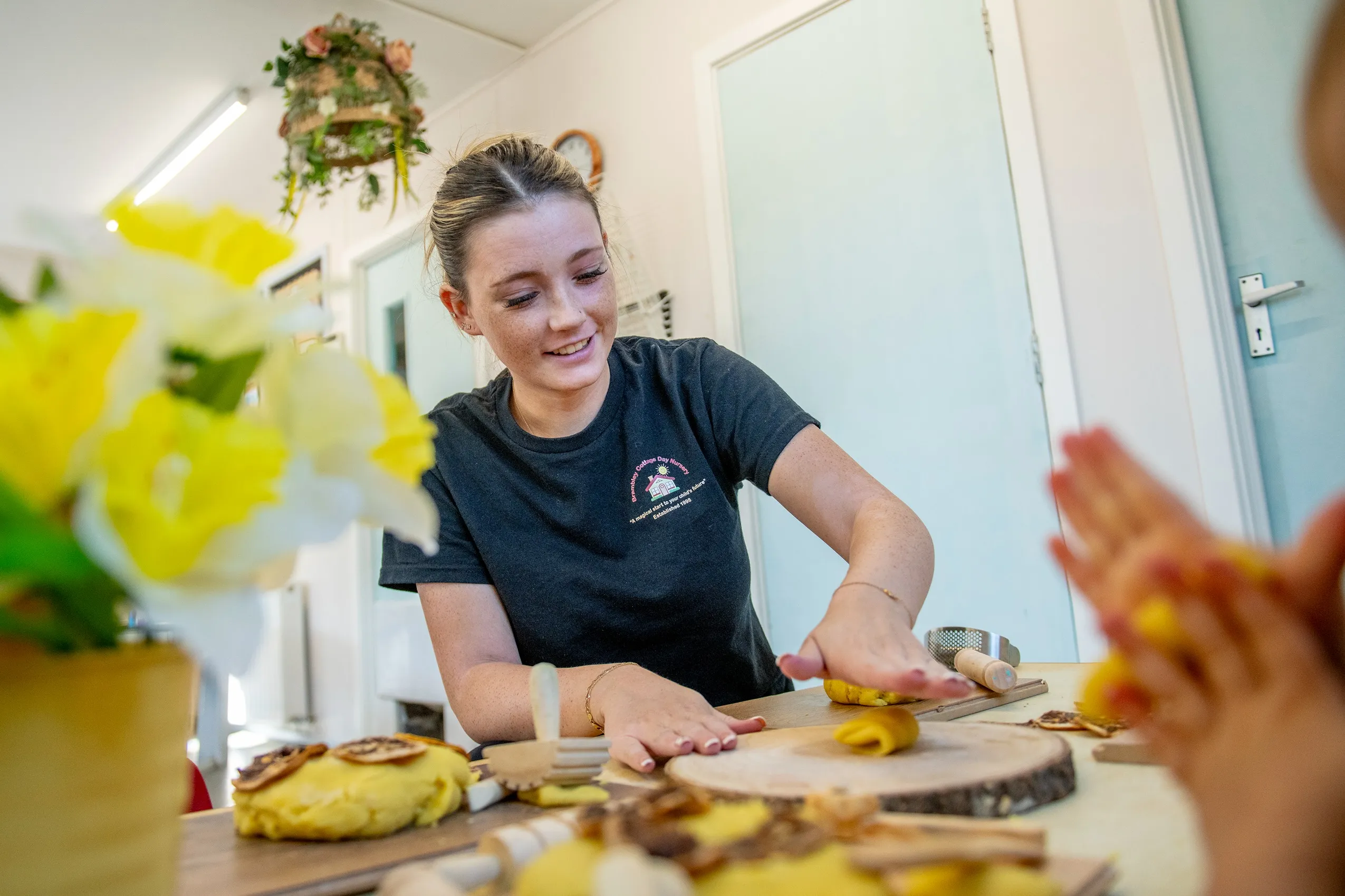 A smiling woman with her hair in a bun, pressing yellow dough on a wooden surface, surrounded by other dough pieces and tools.