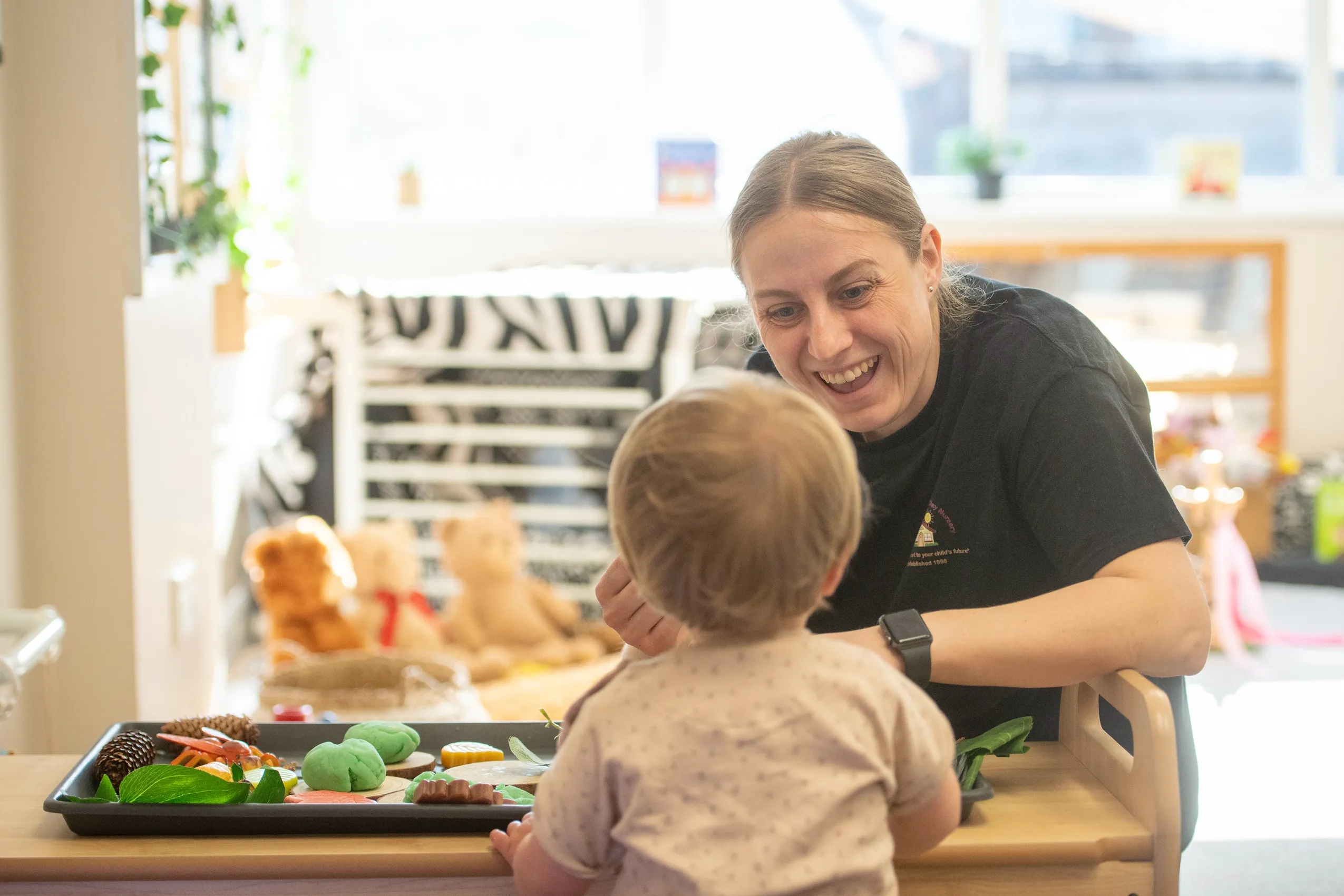 A smiling woman interacting with a young child at a table with a tray of pine cones, leaves, and green playdough.