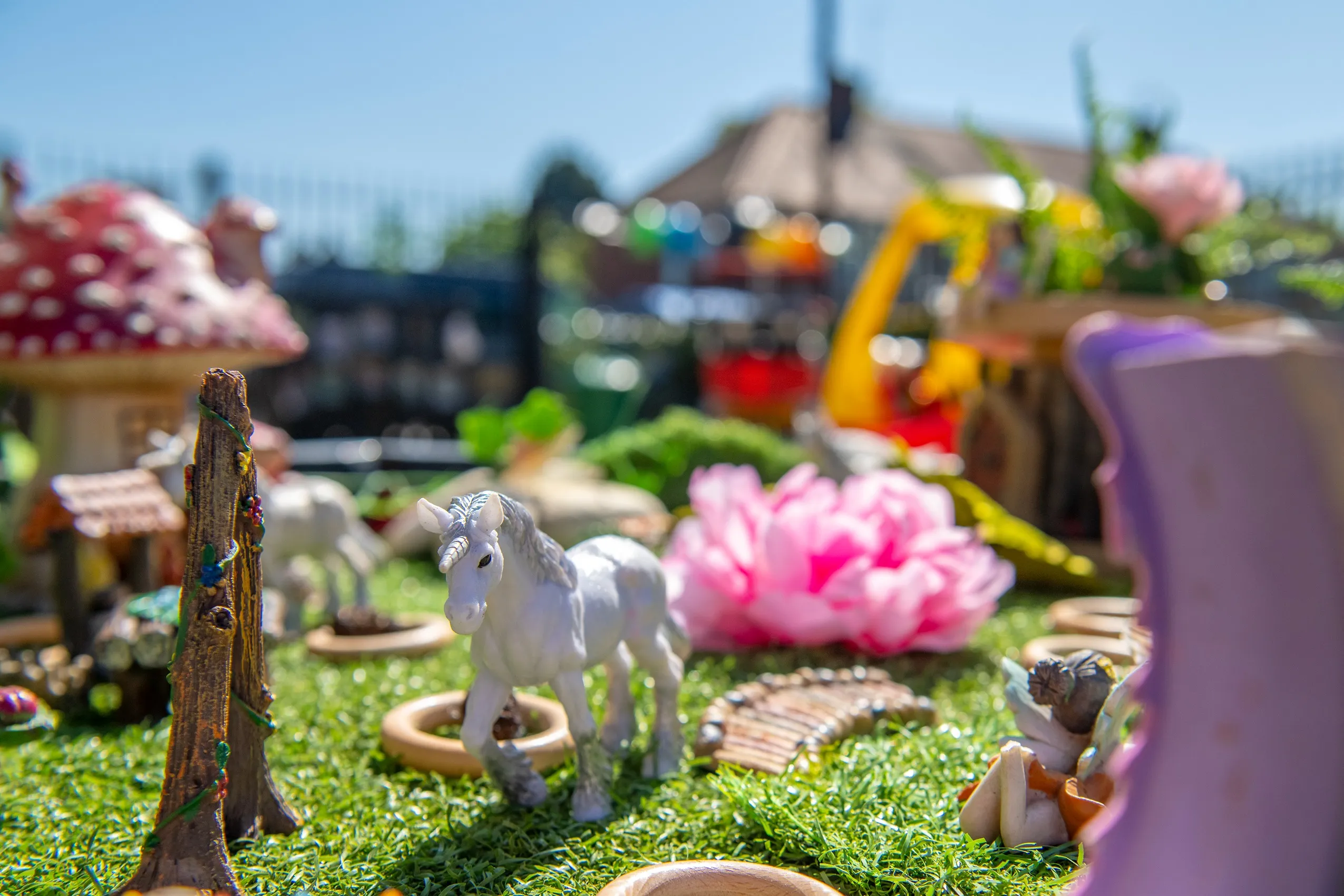 A white unicorn figurine with a gray mane and tail, standing on artificial grass surrounded by miniature fairy garden items.