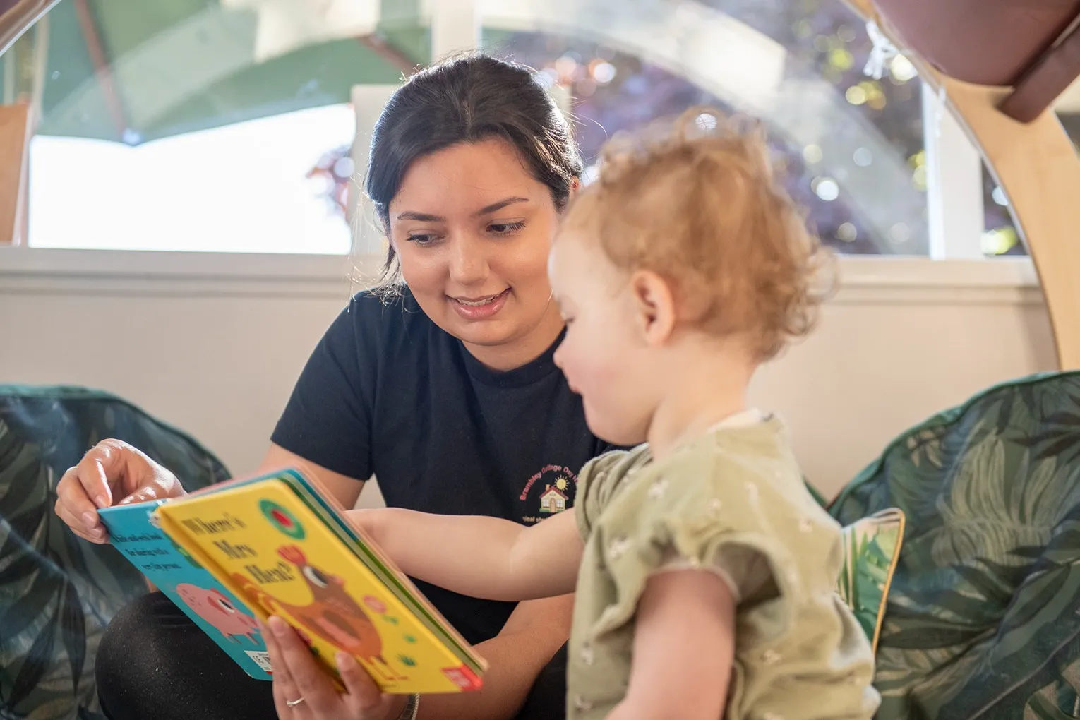 A woman holding a children's book titled 'Where's Mrs Hen?' with a yellow cover and a cartoon bird, as a toddler looks on.