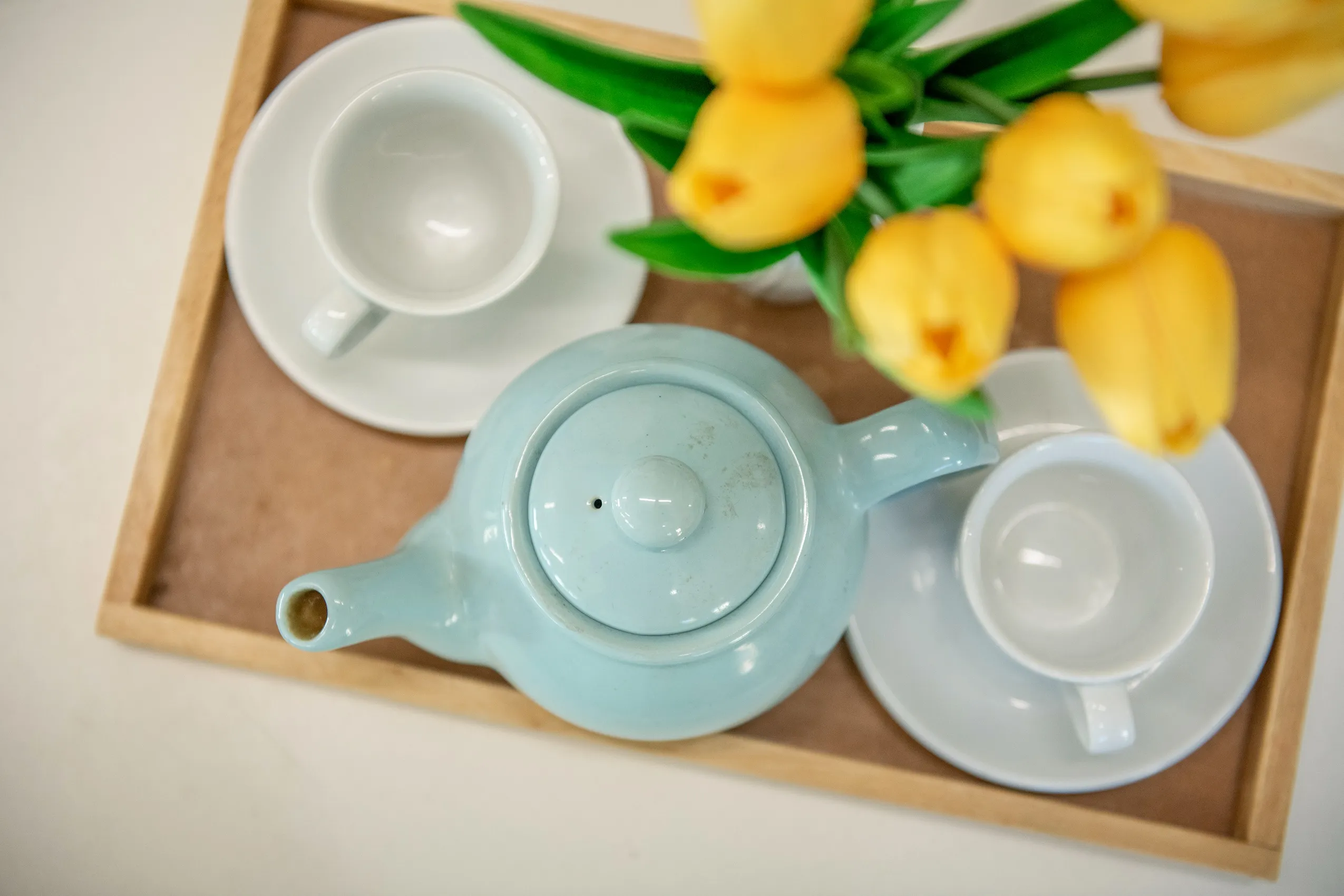 A light blue teapot, two white teacups, and a vase of yellow tulips on a wooden tray.