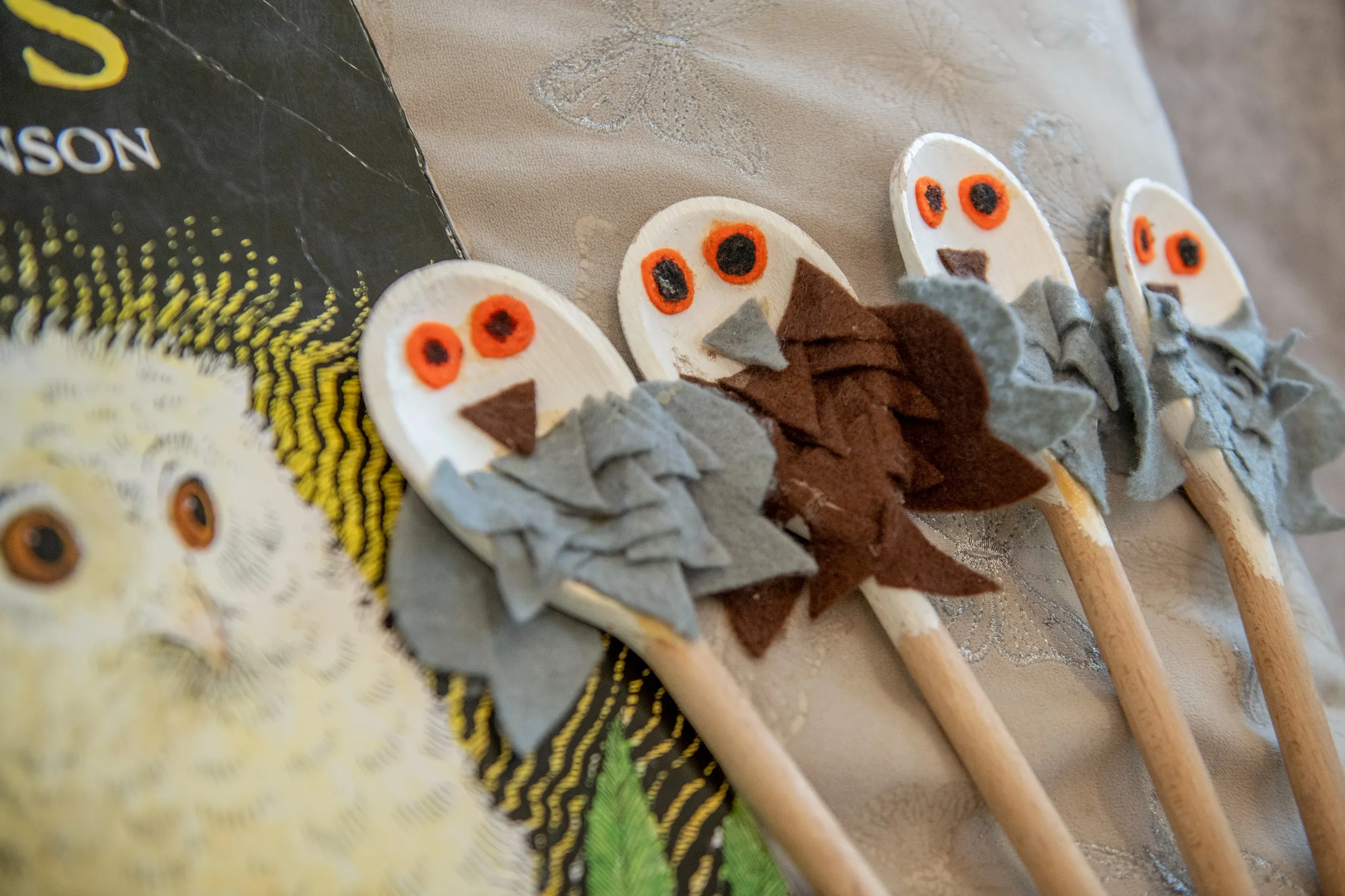 Four wooden spoons decorated as owls with felt wings, next to an owl-themed book.