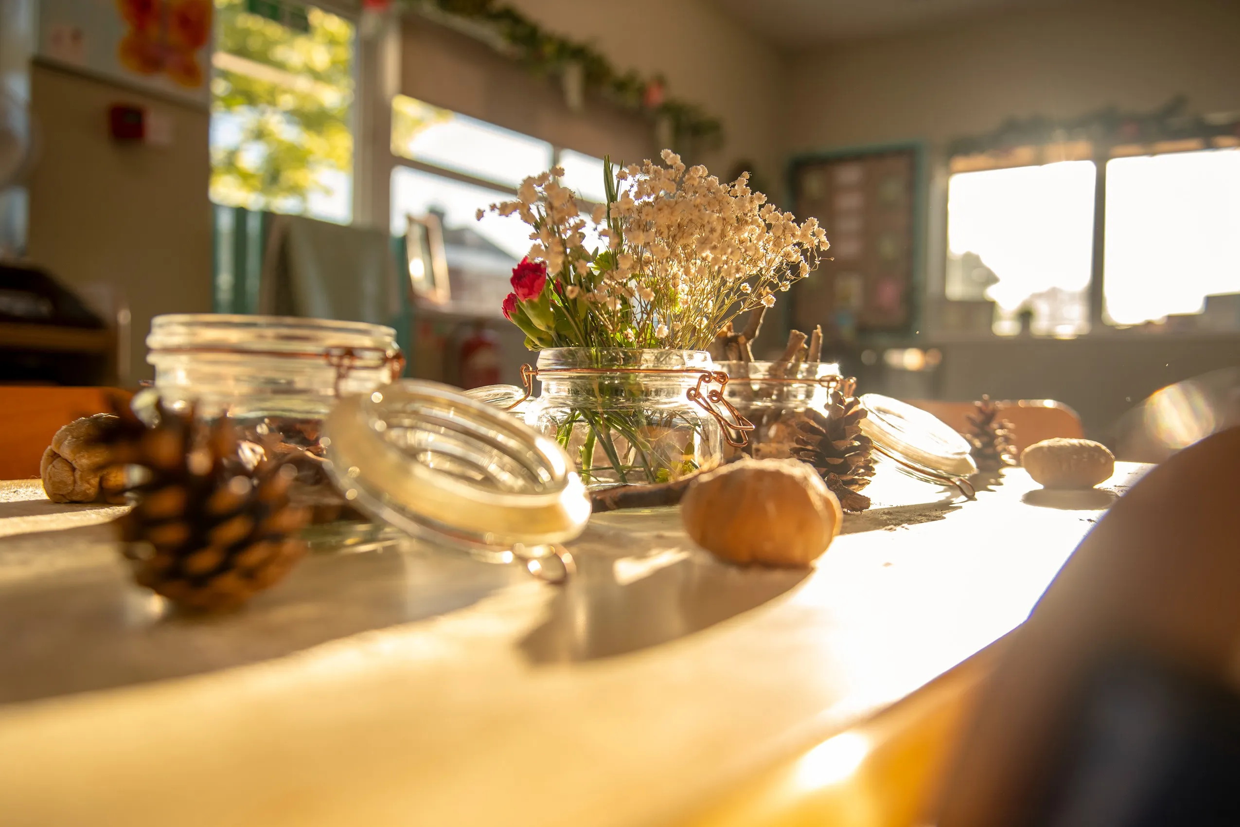 A table arrangement with three glass jars, one with white and red flowers, and scattered pinecones on a wooden table.