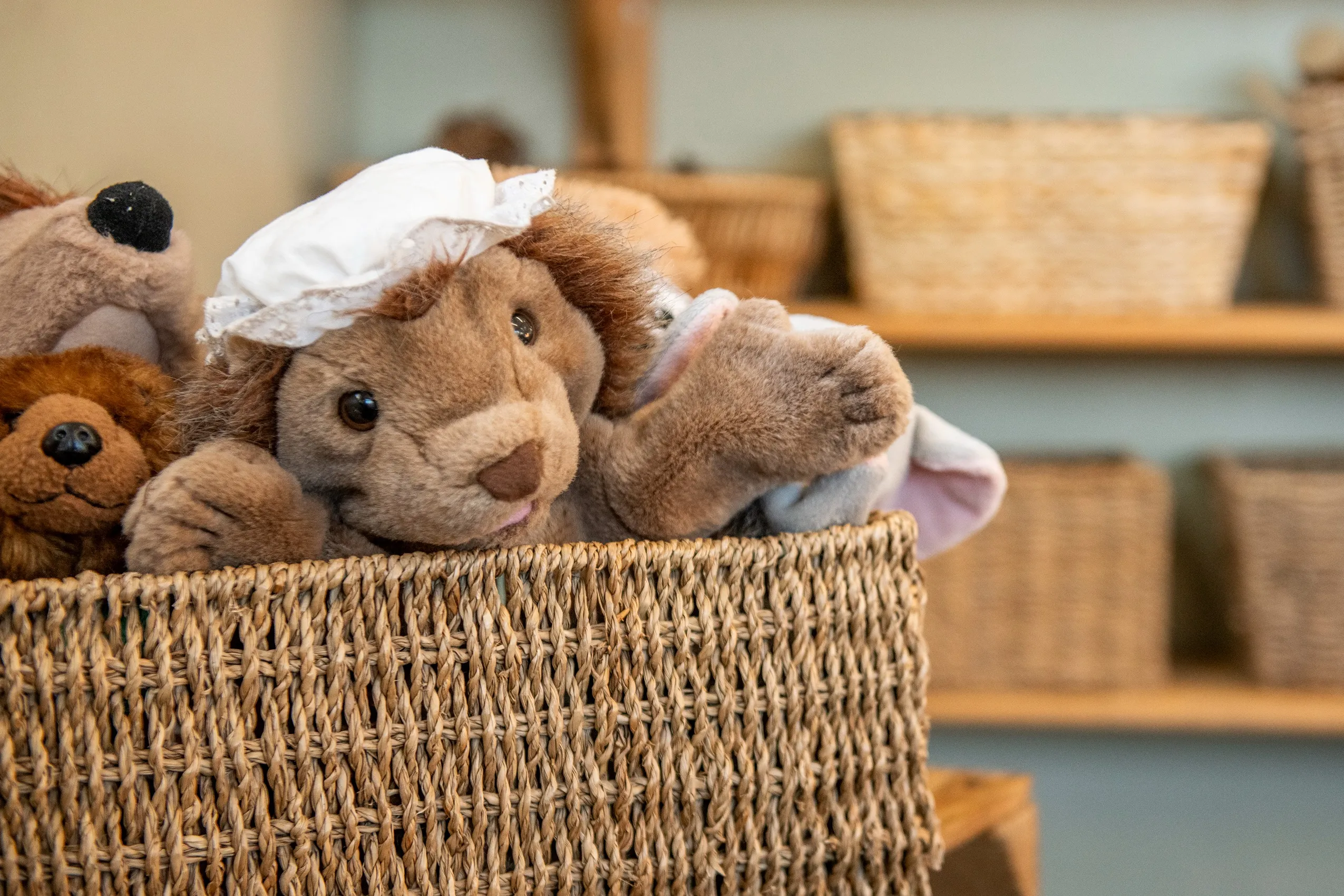 A stuffed lion toy wearing a white bonnet, sitting in a woven basket with other stuffed animals in the background.