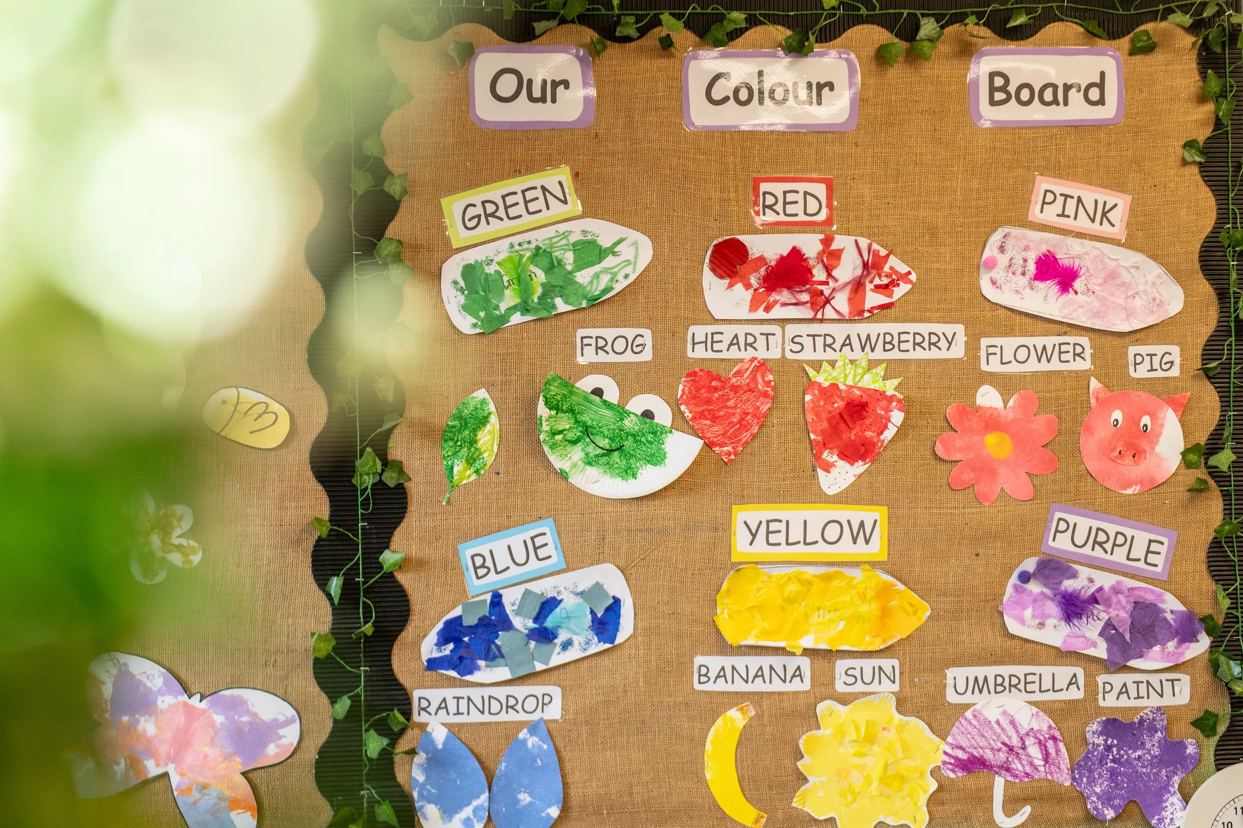 A burlap bulletin board with 'Our Colour Board' signs and rows of colorful children's artwork, each labeled with a color and object.